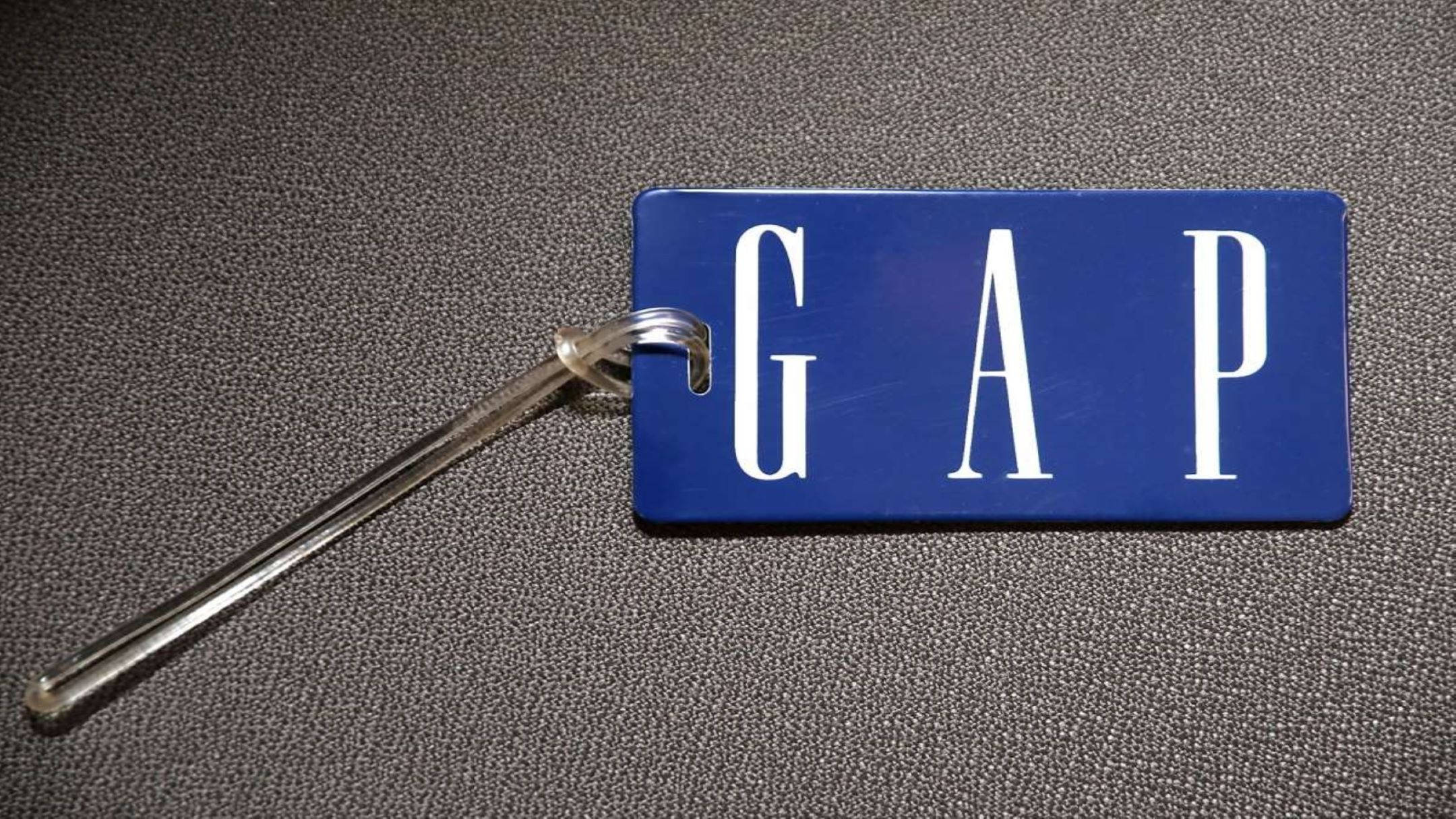 Gap 桌布 壁紙 — 一個藍色Gap行李牌，上面有白色字體，放置在帶有紋理的灰色表面上，並由透明塑料環固定。