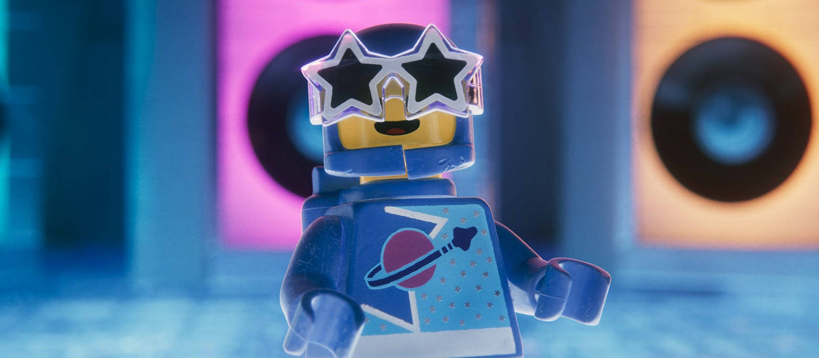 電影動圖 桌布 壁紙 — 一個藍色LEGO人偶戴著星形太陽眼鏡和太空主題襯衫，站在一個色彩繽紛、霓虹燈光、有大喇叭的房間裡。