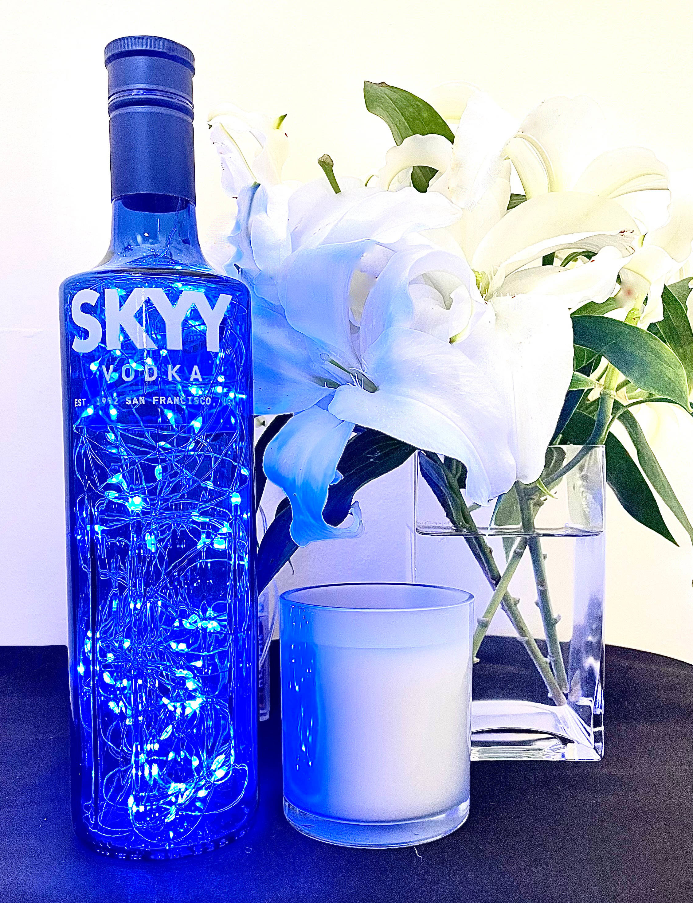 飲料 桌布 壁紙 — 一個藍色Skyy Vodka酒瓶裝滿發光仙女燈，旁邊有白色百合和蠟燭，放在暗色桌子上。