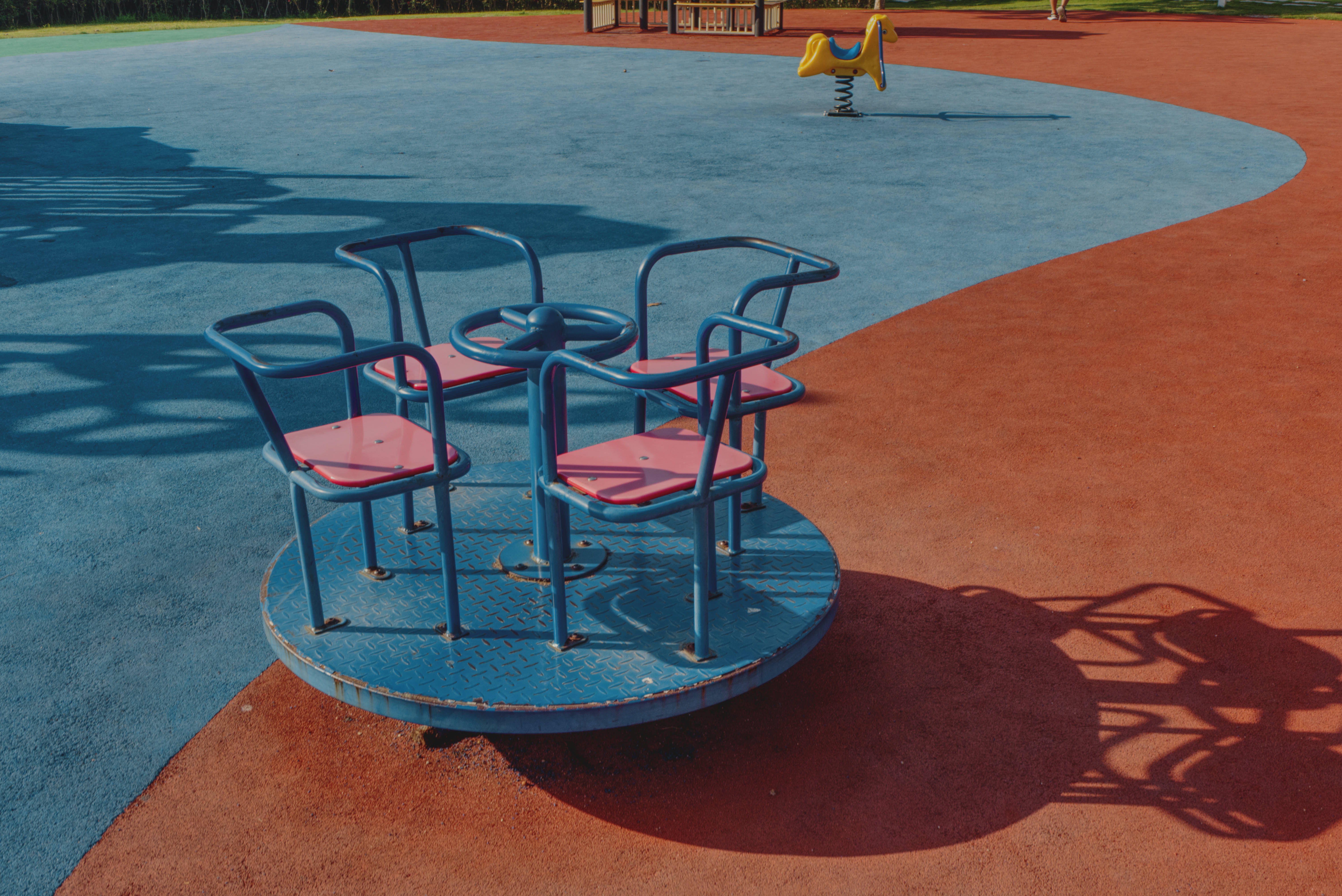 Playground 桌布 壁紙 — 一個藍色和粉紅色的遊樂場旋轉木馬在明亮的陽光下在紅色和藍色的安全表面上投下長長的影子。