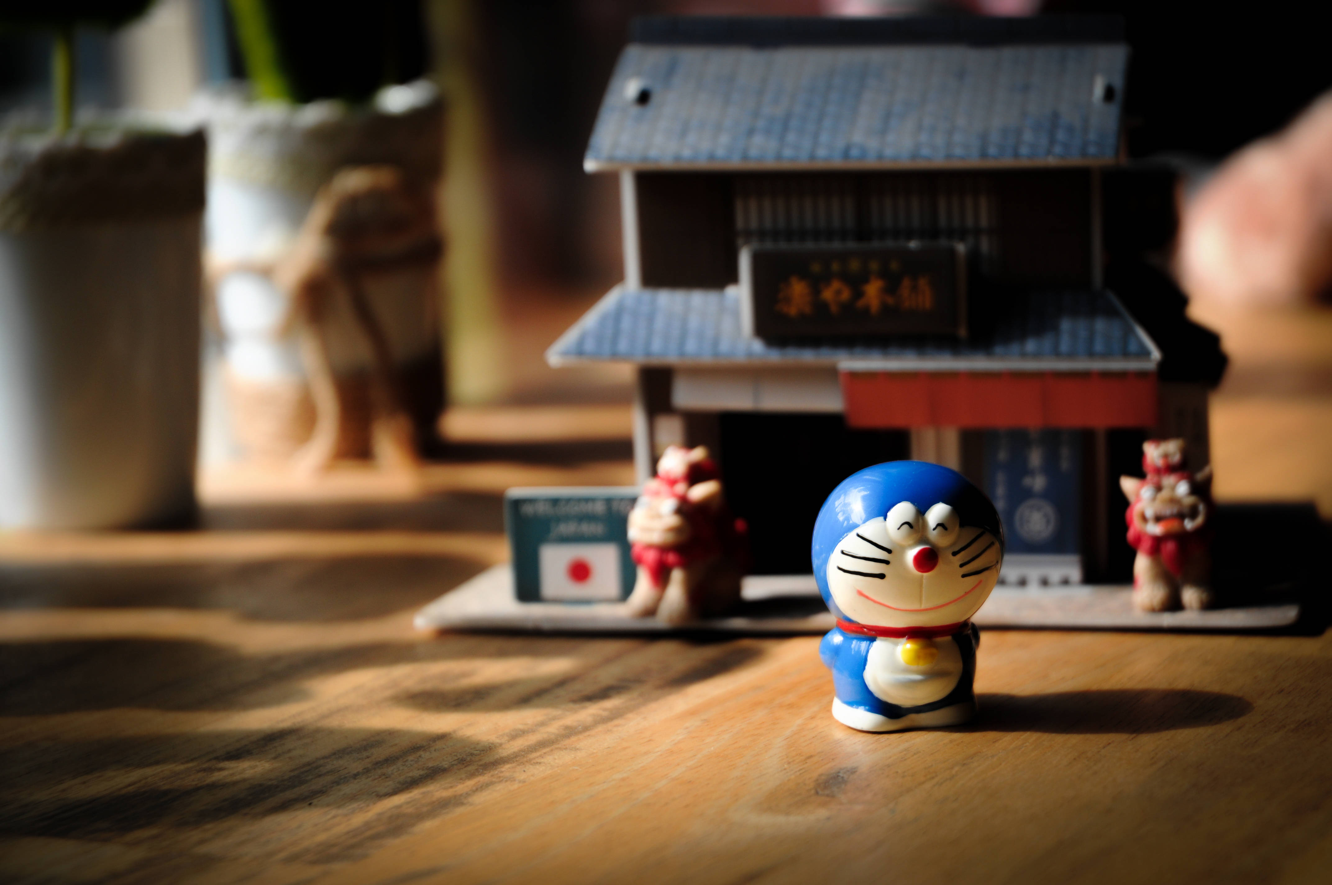 Doraemon 桌布 壁紙 — 一個藍色哆啦A夢公仔坐在木桌上，旁邊有一個帶有紅色燈籠和歡迎牌的微型日本風格房屋。