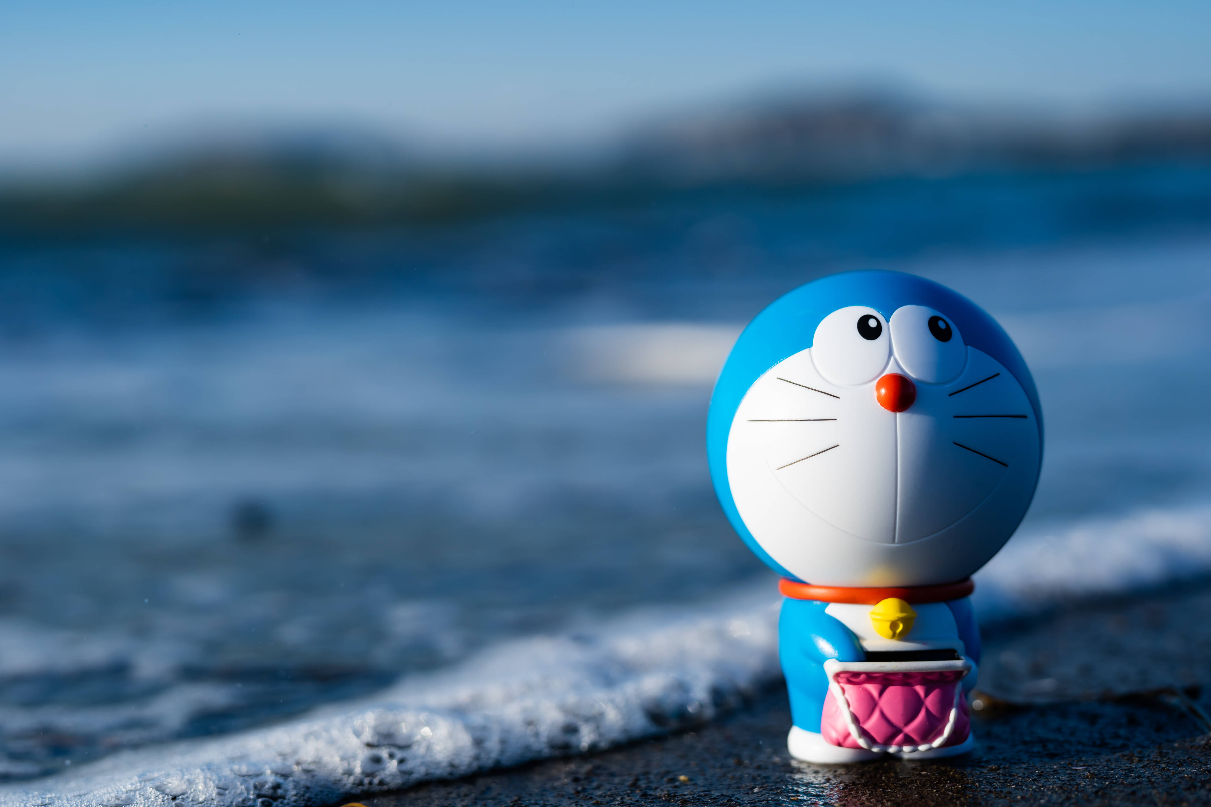 Doraemon 桌布 壁紙 — 一個藍色哆啦A夢公仔站在濕漉漉的沙灘上，溫柔的海浪在晴朗藍天下緩緩湧入。