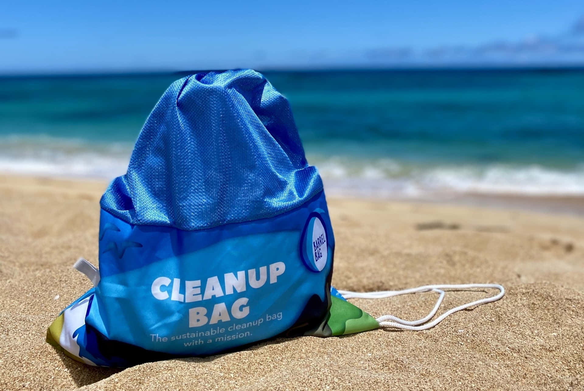 Beach Bag 桌布 壁紙 — 一個藍色束繩清潔袋放在金色沙灘上，碧綠的海浪在清澈的藍天下翻滾而來。