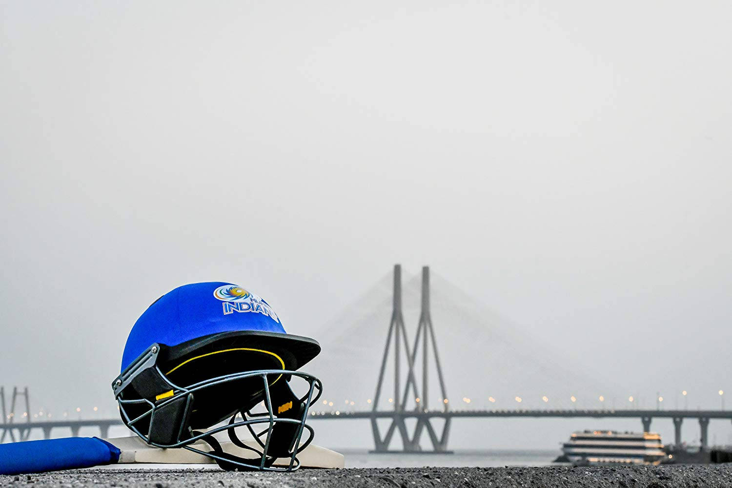 Mumbai Indians 桌布 壁紙 — 一個藍色板球頭盔和球棒放在碼頭上，瓦斯科達伽馬大橋在朦朧的背景中若隱若現。