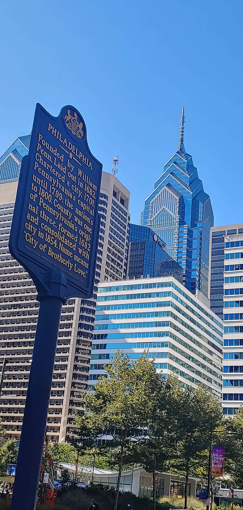 Philly 桌布 壁紙 — 一個藍色歷史標誌矗立在費城現代摩天大樓前，在晴朗的藍天下，下方有綠色樹木。