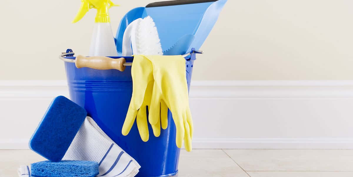 Cleaning 桌布 壁紙 — 一個藍色水桶裝滿黃色手套、海綿、刷子和噴霧瓶，放在瓷磚地板上，靠著白色牆壁。