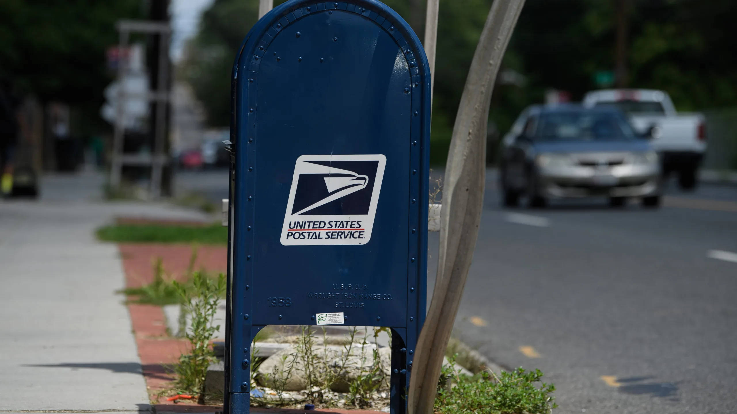 Usps 桌布 壁紙 — 一個藍色的美國郵政信箱放在人行道上，旁邊有樹，車輛在陽光明媚的街道上行駛。
