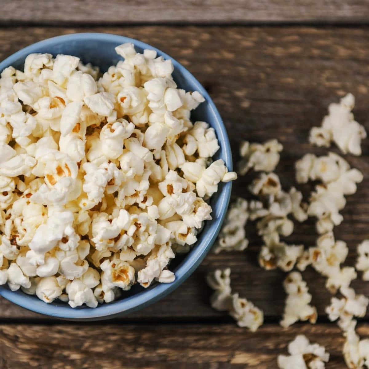 Popcorn 桌布 壁紙 — 一個藍色碗裝滿了蓬鬆的白色爆米花，放在質樸的木桌上，有玉米粒灑出。