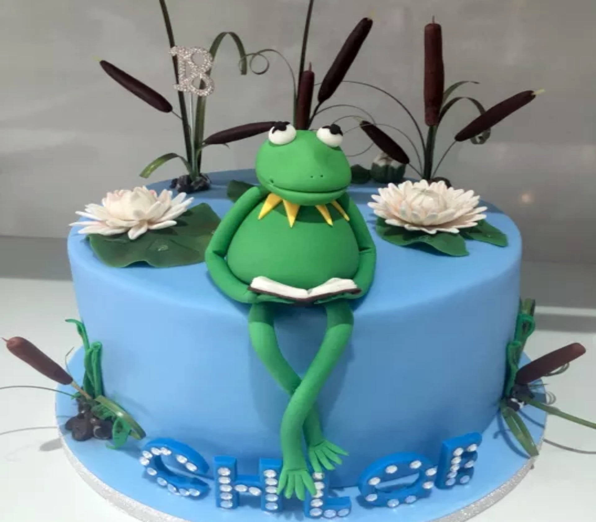 kermit 桌布 壁紙 — 一個藍色糖霜蛋糕上有一個綠色青蛙公仔正在讀書，周圍環繞著白色睡蓮和香蒲。