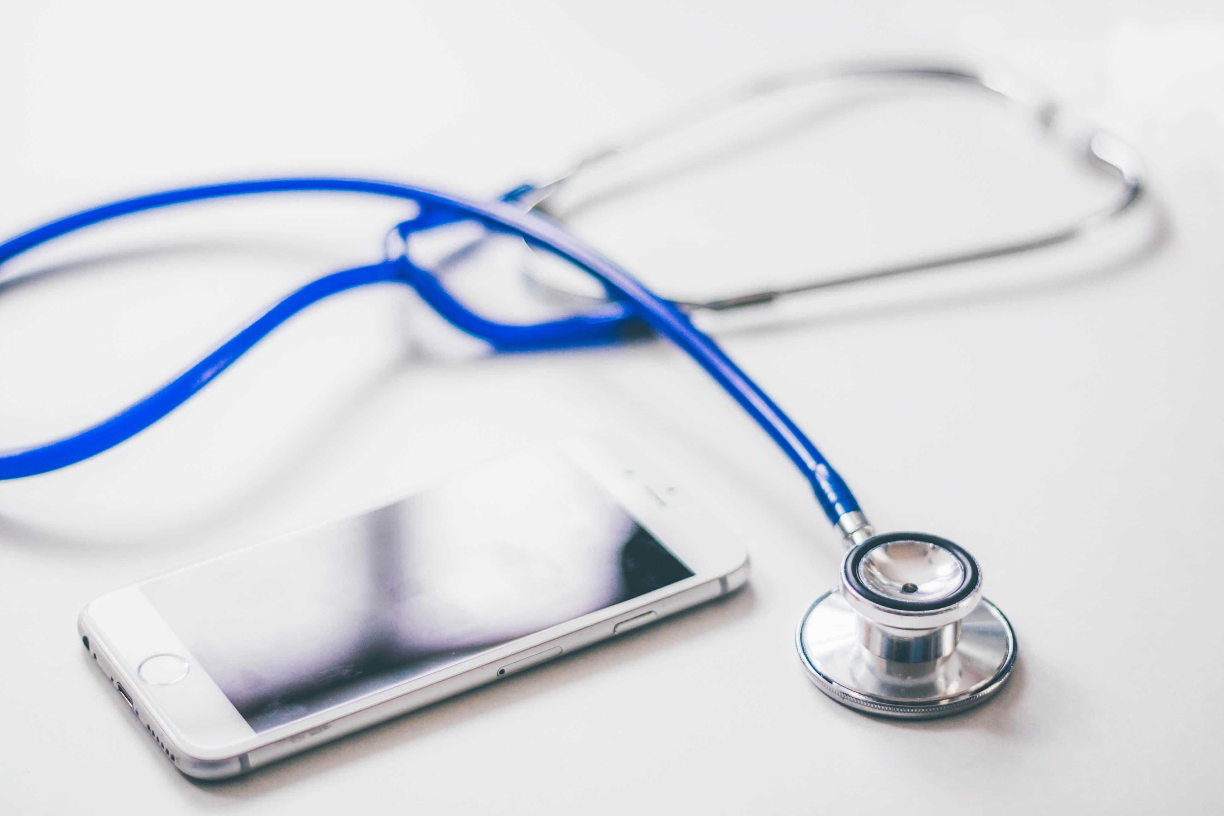Stethoscope 桌布 壁紙 — 一個藍色聽診器放在一支白色智慧型手機旁邊，置於潔白表面上，帶有柔和陰影。