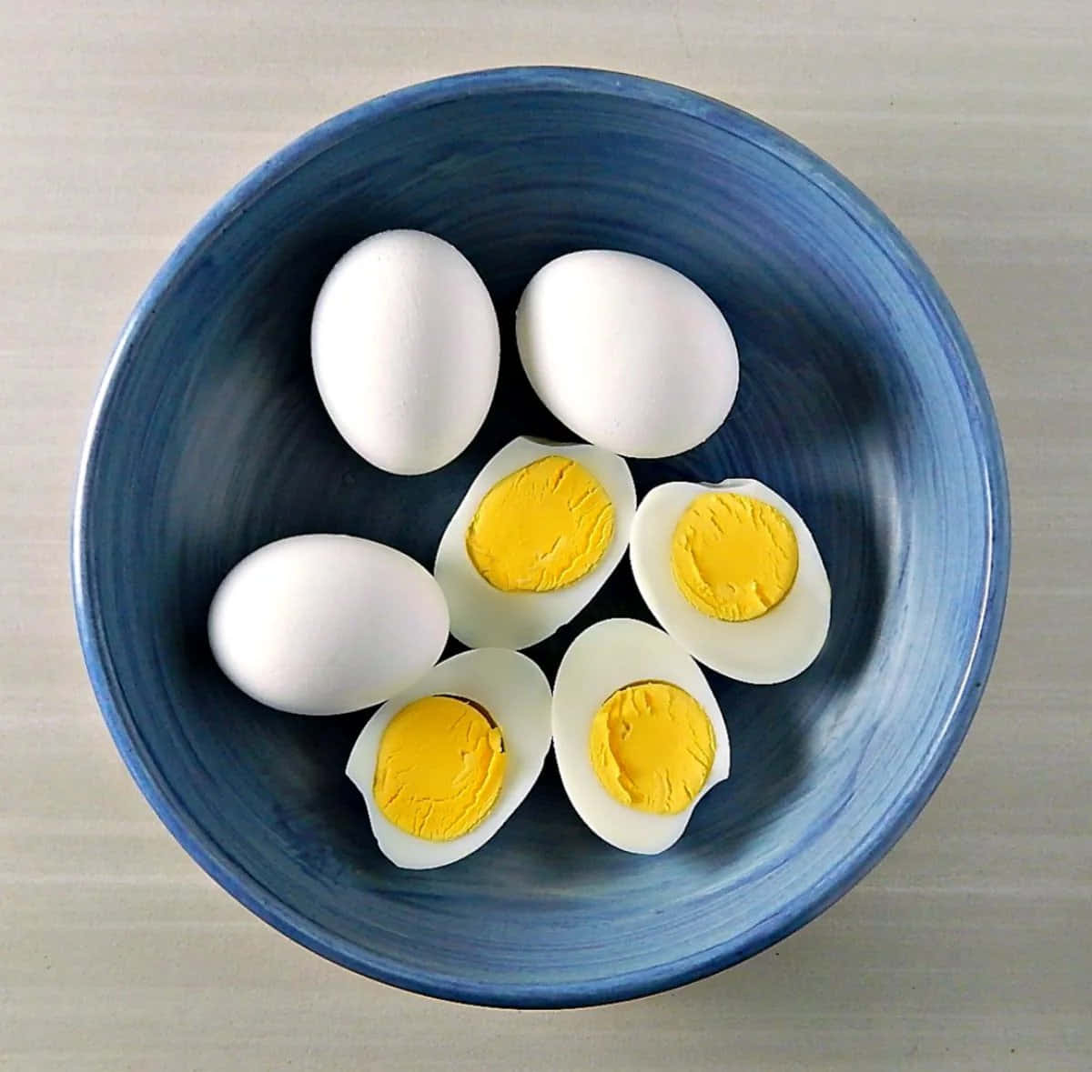 Egg 桌布 壁紙 — 一個藍色陶瓷碗裝著六個水煮蛋，其中三個切半，露出鮮黃色的蛋黃。
