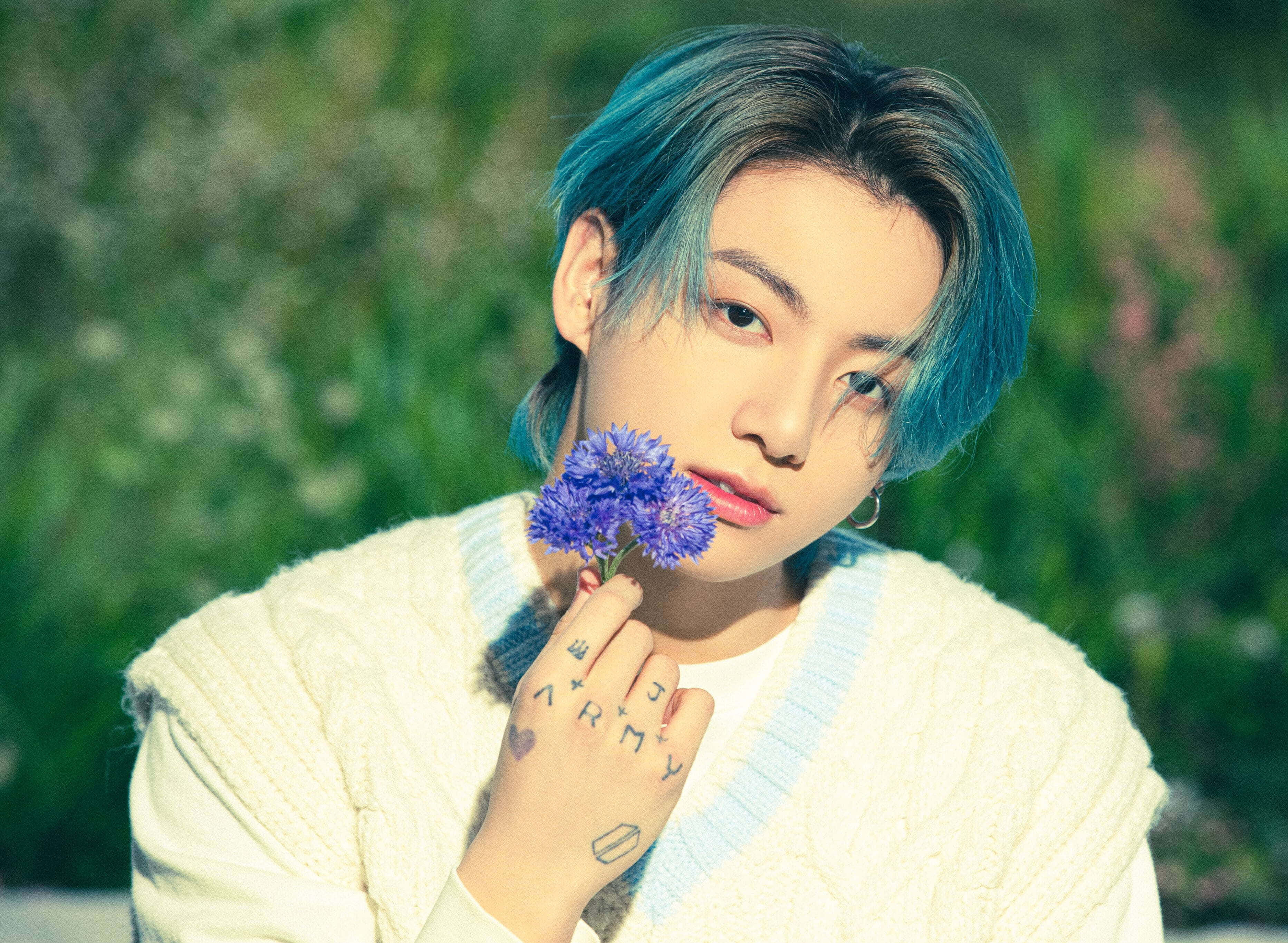 Jungkook 桌布 壁紙 — 一個藍色頭髮的年輕人，穿著奶油色毛衣，在模糊的綠色背景前拿著一朵紫色花。