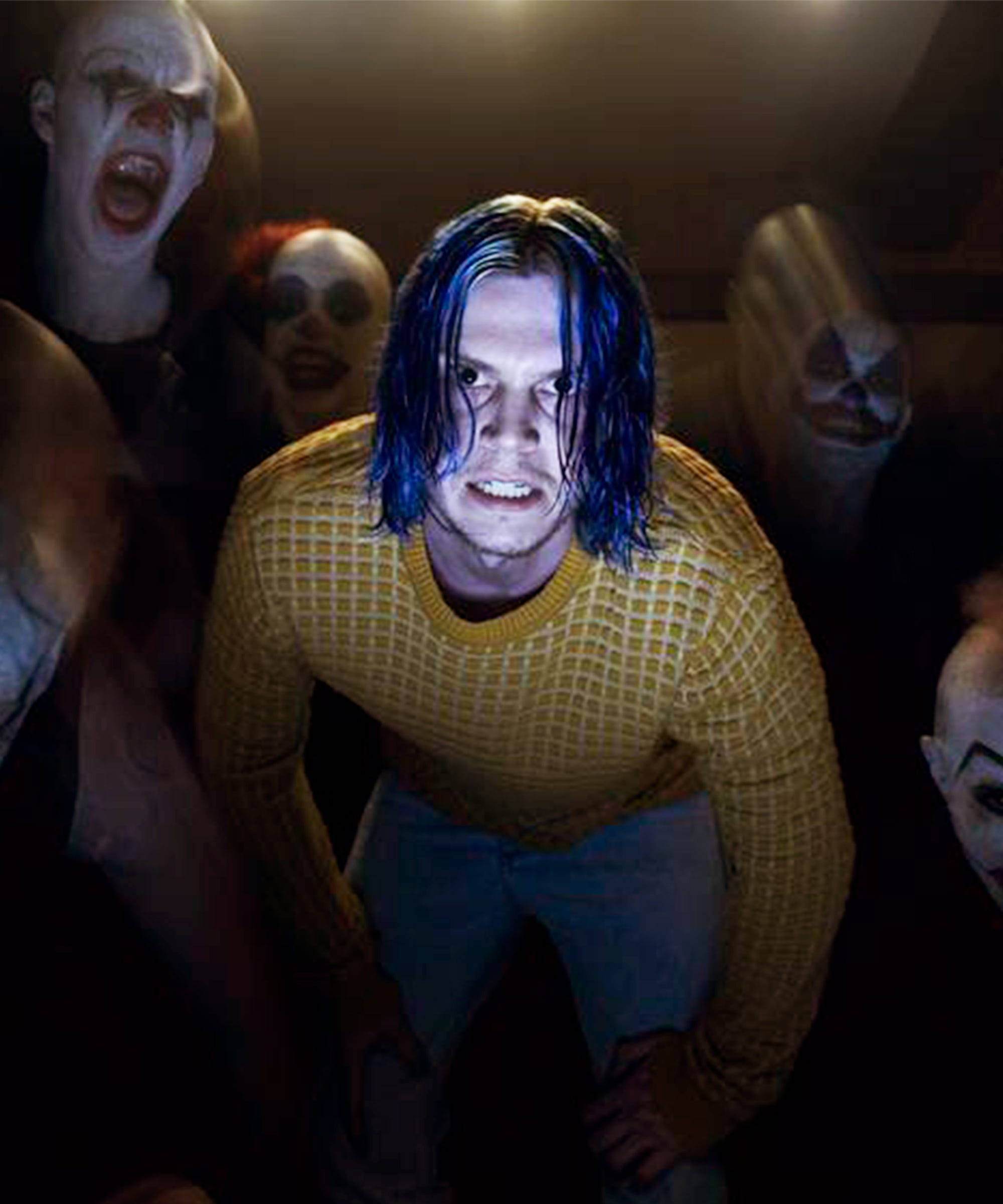 Tate Langdon 桌布 壁紙 — 一個藍色頭髮的驚恐男子蹲在一個黑暗的房間裡，周圍是令人恐懼的小丑人偶。