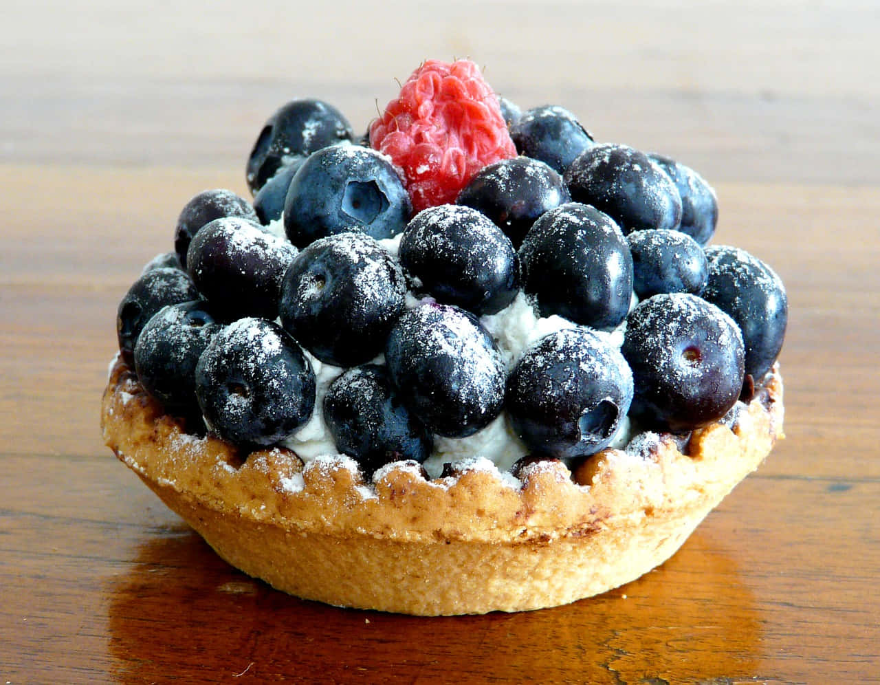 Blueberries Tart 桌布 壁紙 — 一個藍莓塔的近距離特寫，上面撒有糖粉和單粒覆盆子，放在木桌上。