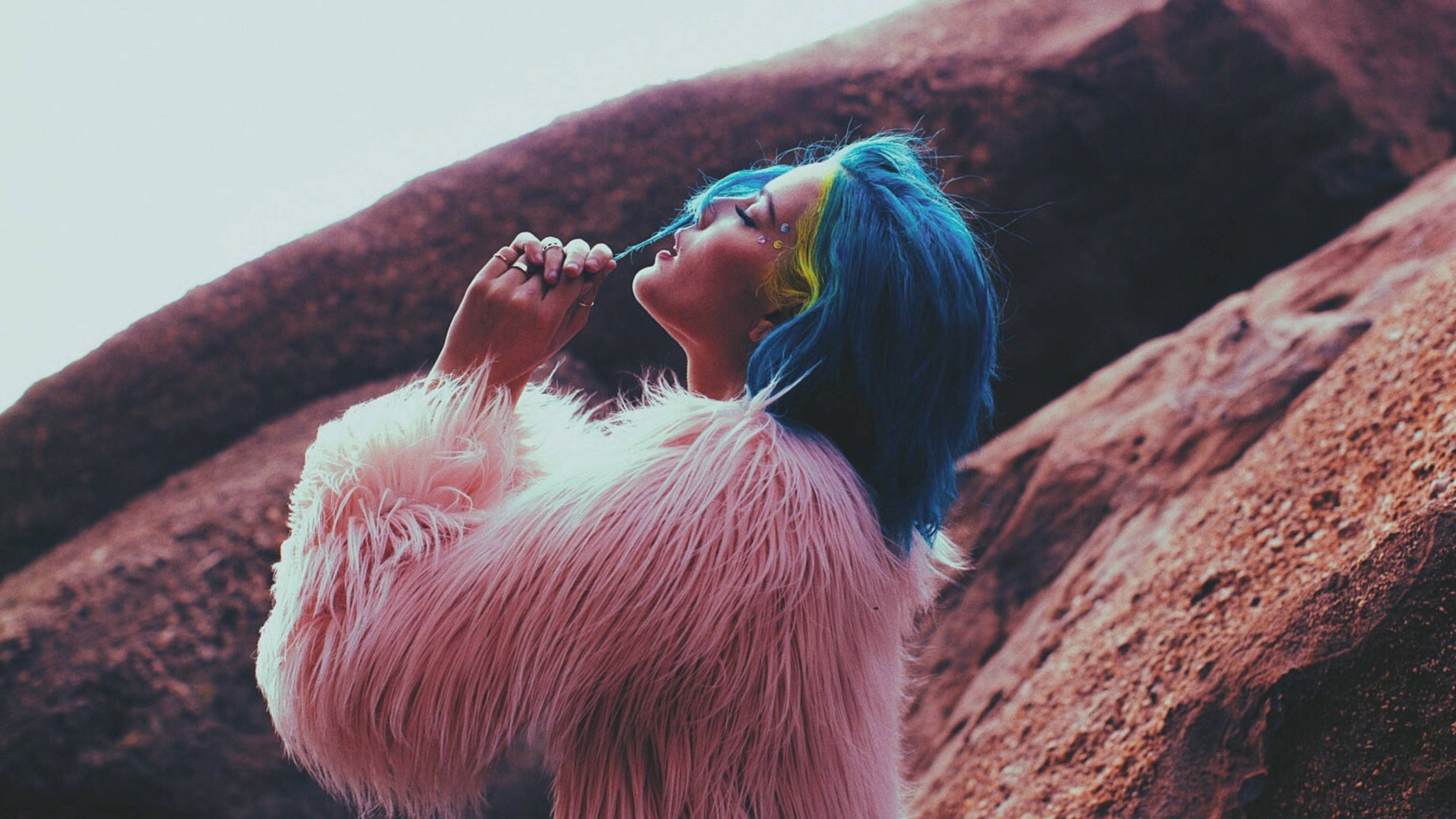 Halsey 桌布 壁紙 — 一個藍黃色頭髮的人穿著毛茸茸的粉紅色外套，在微紅色的岩層背景下吸煙。