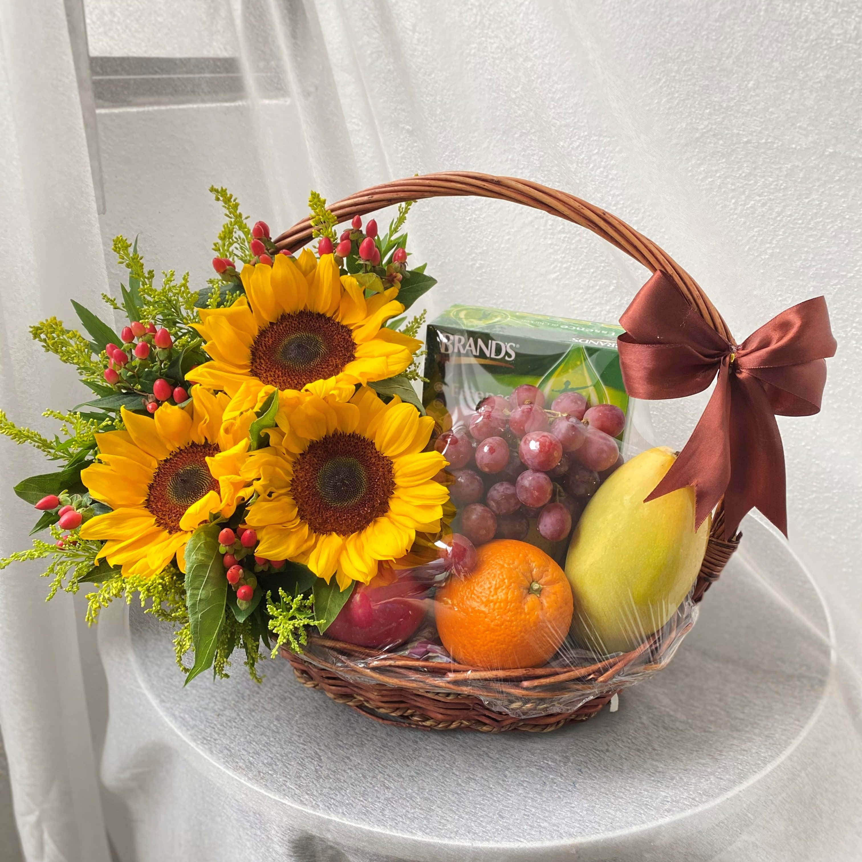 水果籃 桌布 壁紙 — 一個藤籃裝滿明亮的向日葵、葡萄、芒果和柳橙，放在一張白色桌子上，繫著紅色蝴蝶結。