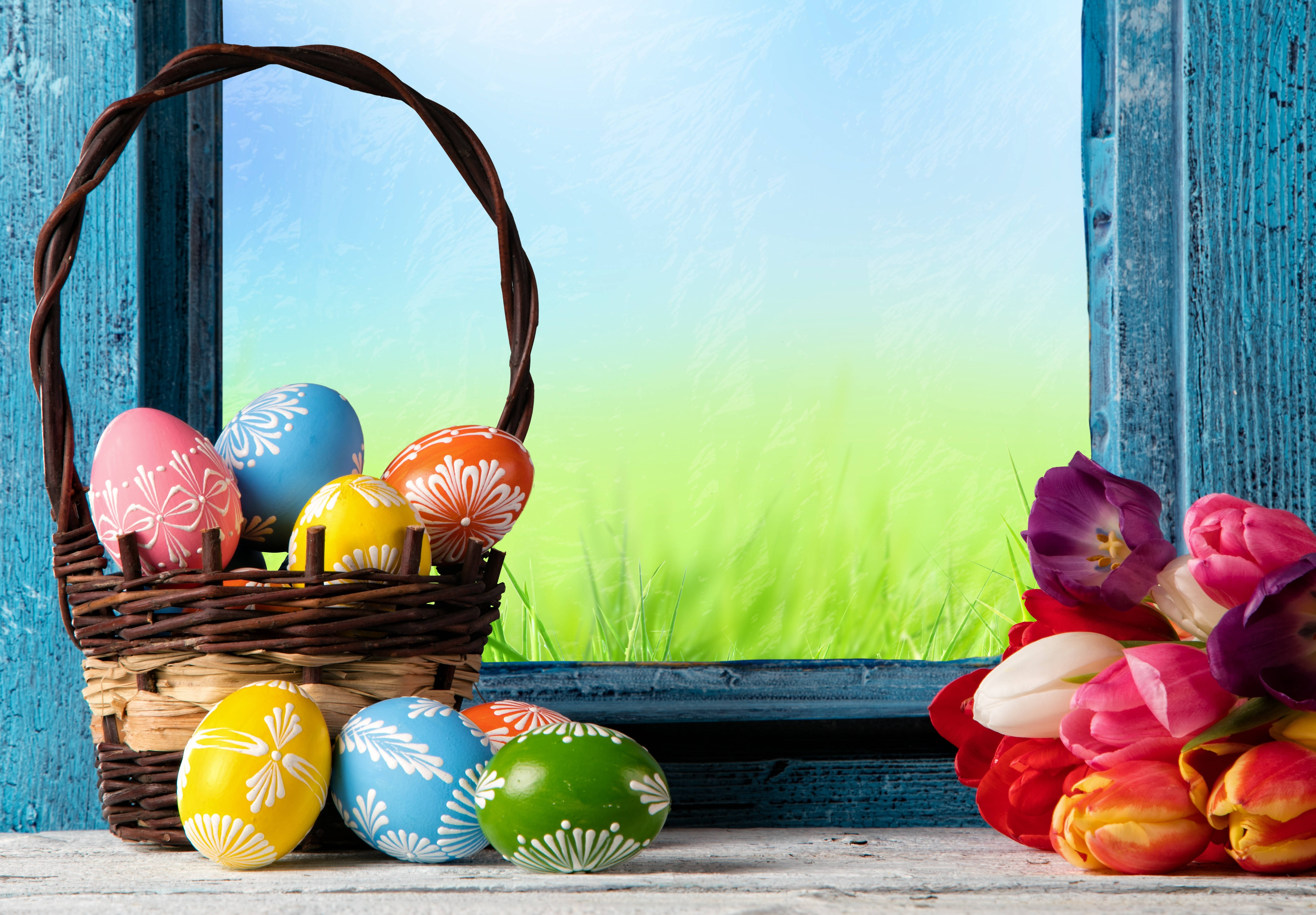 Cute Easter 桌布 壁紙 — 一個藤籃裝滿色彩繽紛的圖案復活節蛋，放在窗台上，旁邊是一束鮮豔的鬱金香，窗外是綠草如茵。