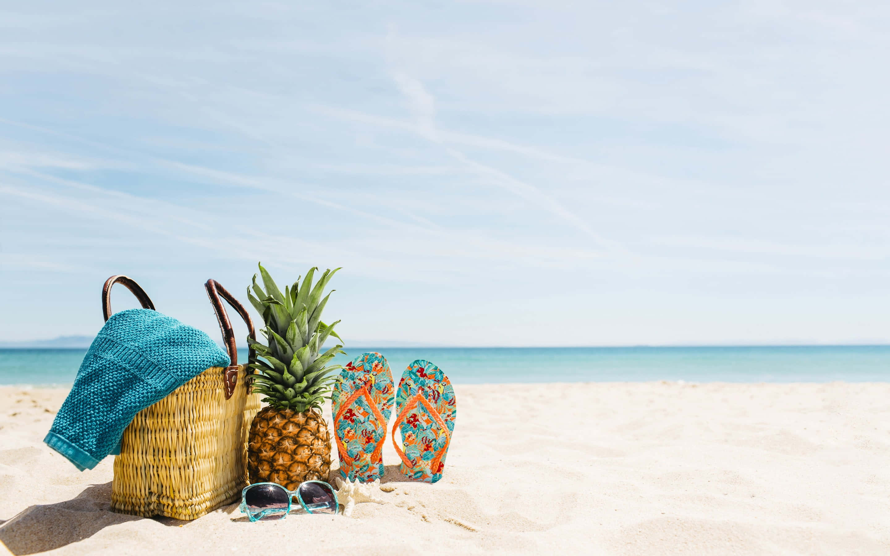 Beach Bag 桌布 壁紙 — 一個藤編沙灘袋、藍綠色毛巾、鳳梨人字拖和太陽眼鏡放在陽光照耀的沙灘上，旁邊是平靜的藍綠色海洋。