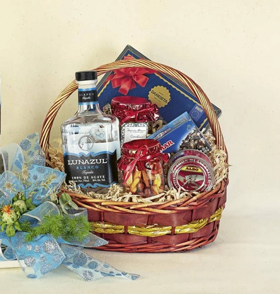 alcohol 桌布 壁紙 — 一個藤編禮籃，裝滿龍舌蘭酒、美食小吃和藍絲帶花束，靠著中性色牆壁。