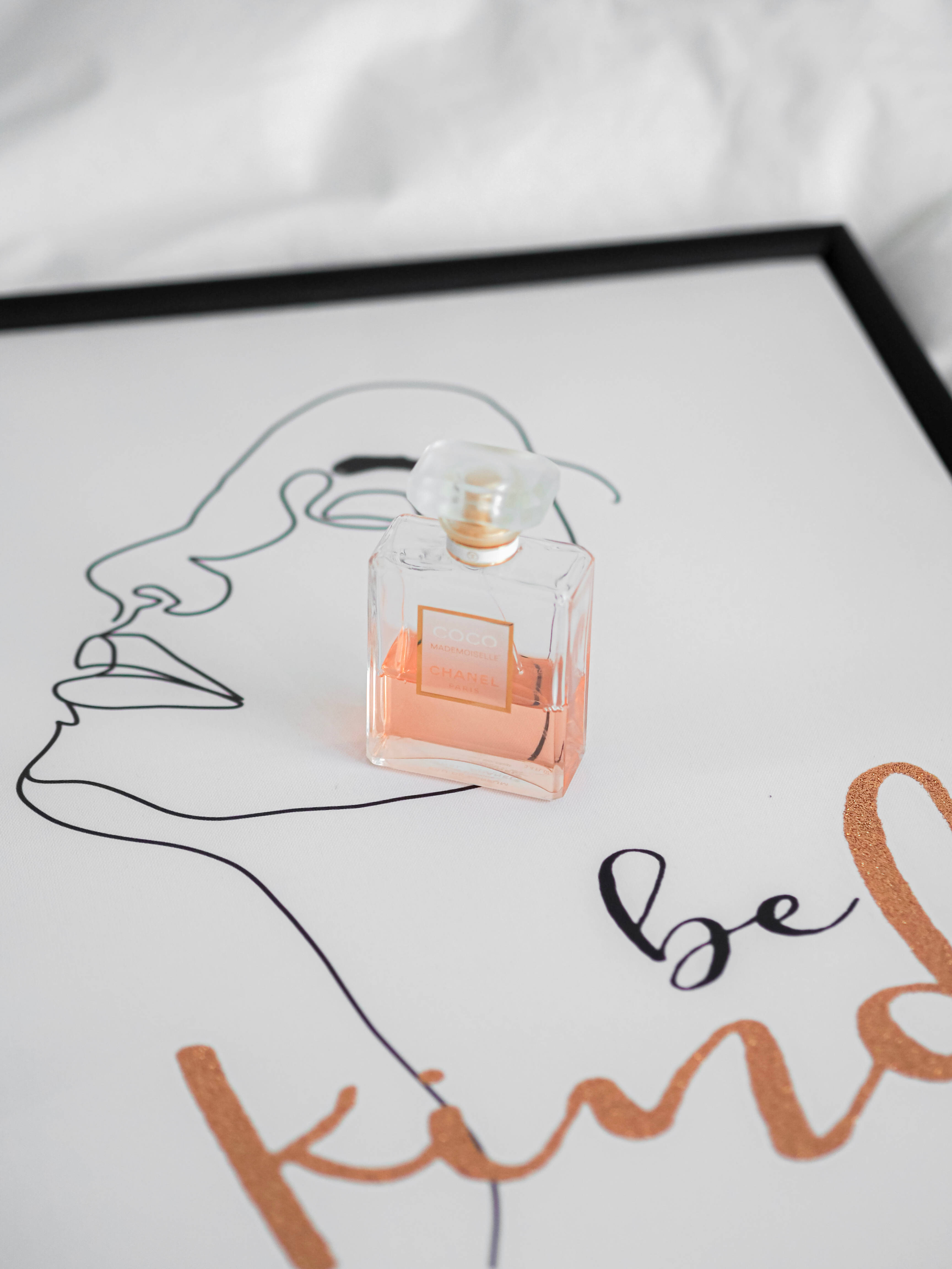 Chanel 桌布 壁紙 — 一個蜜桃香水瓶放在一個簡約的裱框藝術印刷品上，上面有線條畫和「be kind」文字。