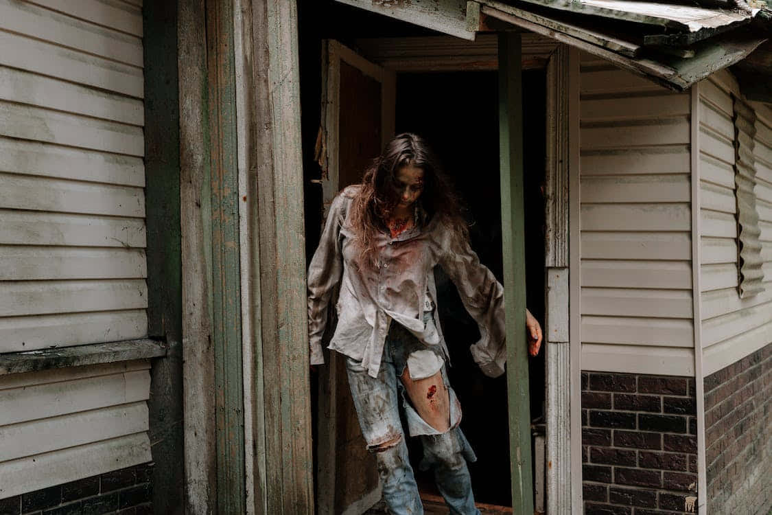 Scary Zombie 桌布 壁紙 — 一個血淋淋、頭髮蓬亂的殭屍從破舊的房屋門口出現，周圍是破敗的郊區環境。