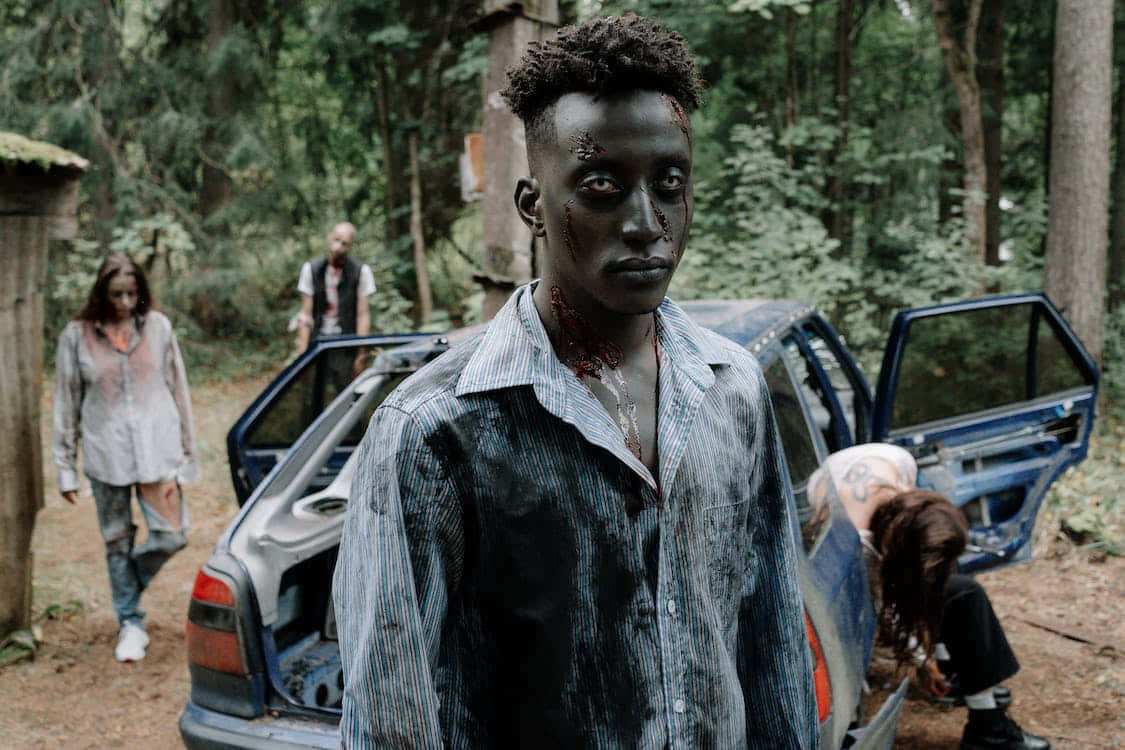 Scary Zombie 桌布 壁紙 — 一個血淋淋的殭屍在森林空地中，旁邊有一輛破爛的藍色汽車，附近還有其他不死生物，牠正凝視著前方。