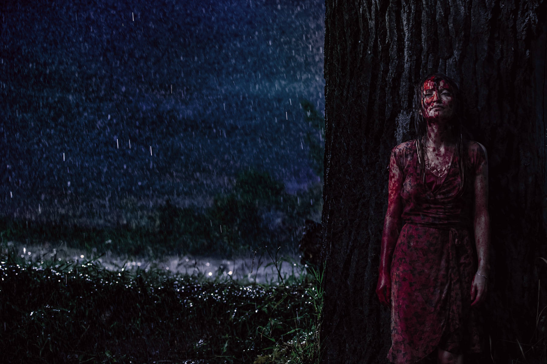 Gods 桌布 壁紙 — 一個血淋淋的身影在夜間暴雨中靠著樹幹一動不動，背景是深藍色的天空和發光的濕草。