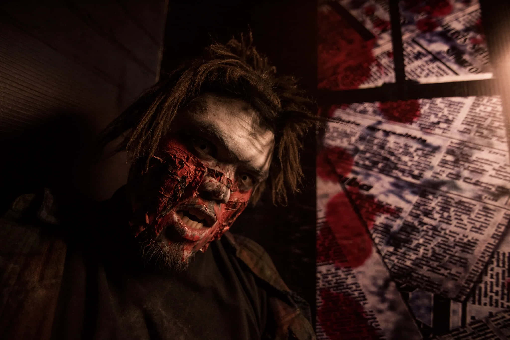 Scary Zombie 桌布 壁紙 — 一個血腥的殭屍，臉上沾滿血，在燈光昏暗的房間裡，牆上還有染血的報紙，牠正狠狠地盯著。