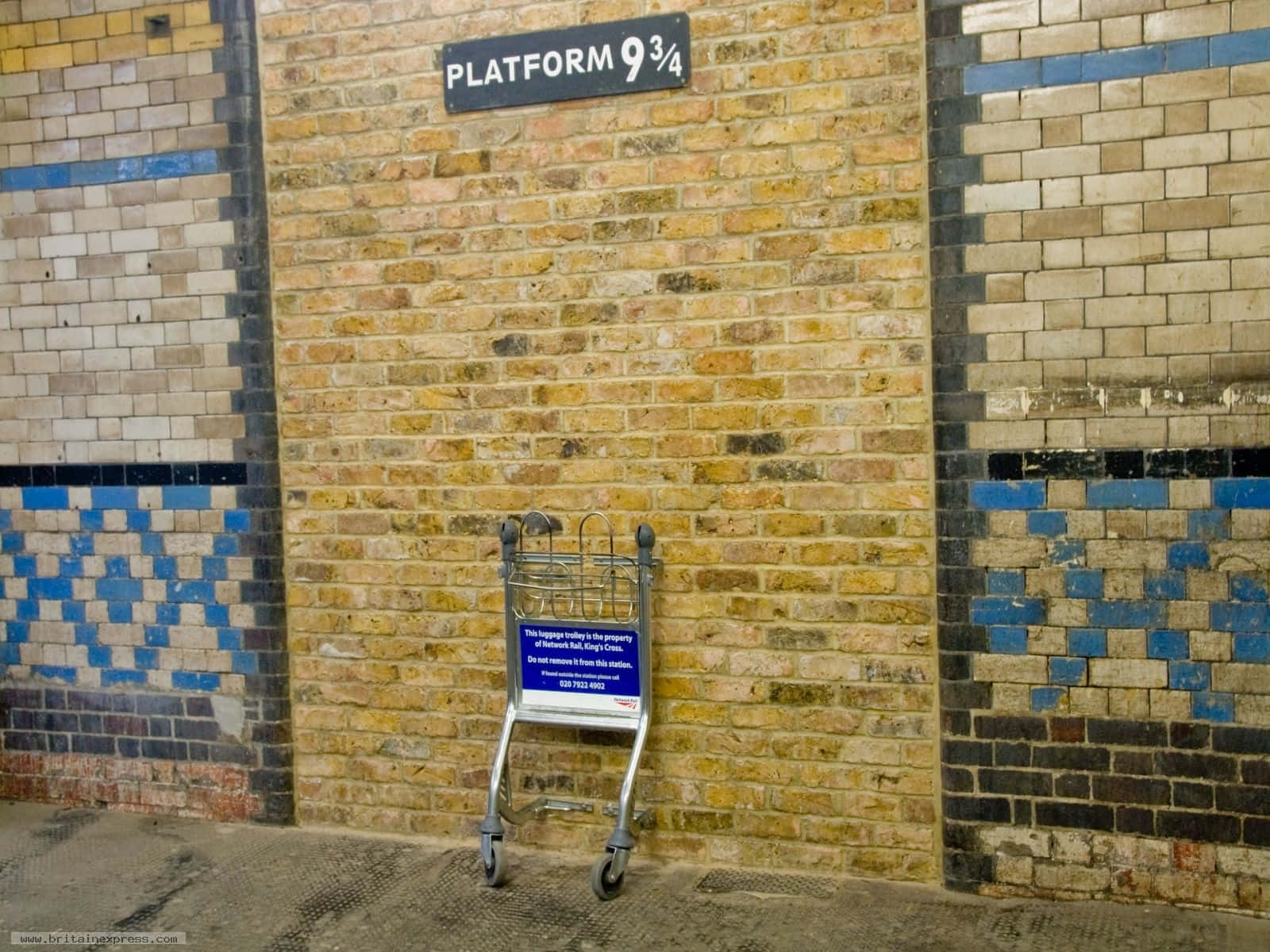 Platform 9 3/4 桌布 壁紙 — 一個行李手推車靠在火車站的磚牆上，在9¾月台標誌下方。