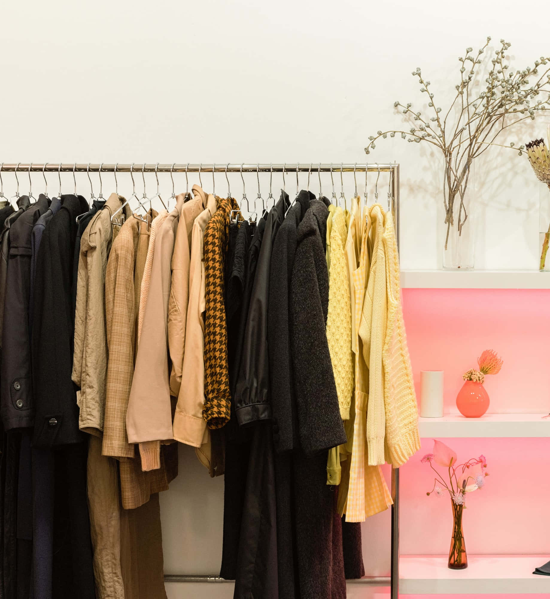 Clothes 桌布 壁紙 — 一個衣架上展示了一系列精選的大衣和毛衣，色調以大地色和亮黃色為主，旁邊是粉紅色燈光的層架，擺放著乾花裝飾。