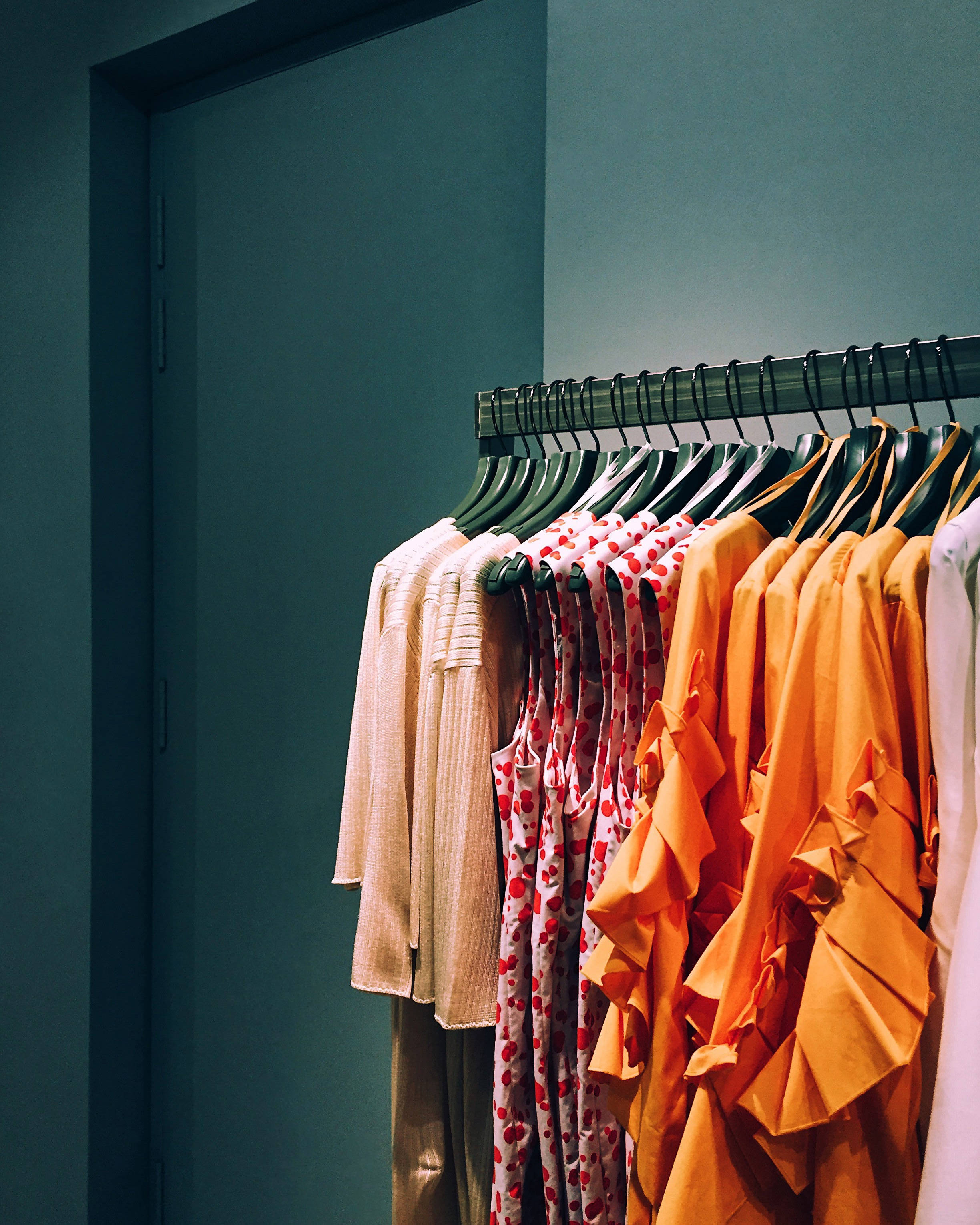 Trendy 桌布 壁紙 — 一個衣架展示色彩繽紛的衣物，背景是藍綠色牆壁和一個黑暗的門口。