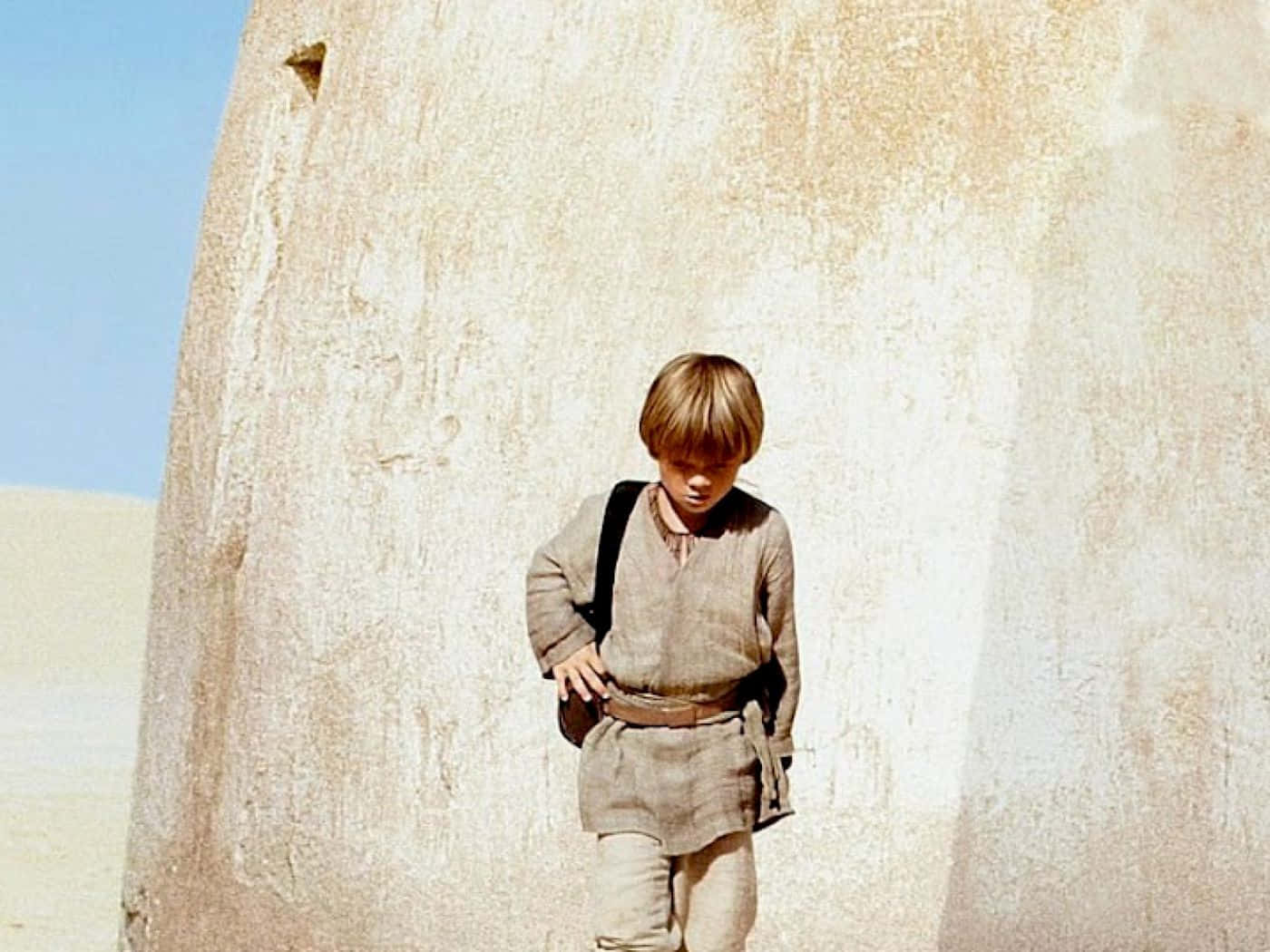 The Phantom Menace 桌布 壁紙 — 一個衣衫襤褸的小男孩在晴朗的藍天下，莊嚴地站在一個巨大的風化石頭結構前。