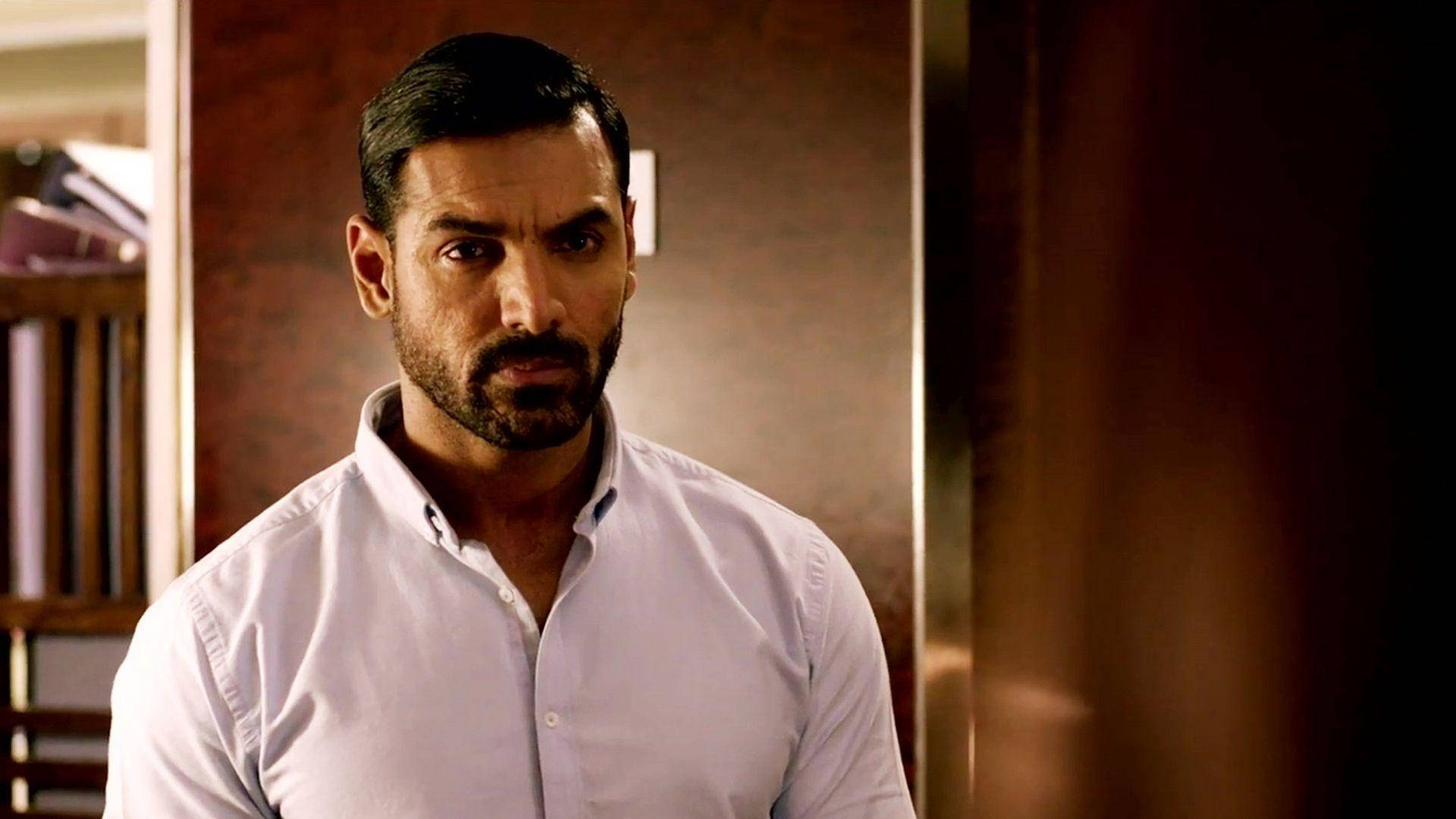 John Abraham 桌布 壁紙 — 一個表情嚴肅的男人在溫暖光線的室內環境中向前凝視，身後有木架。