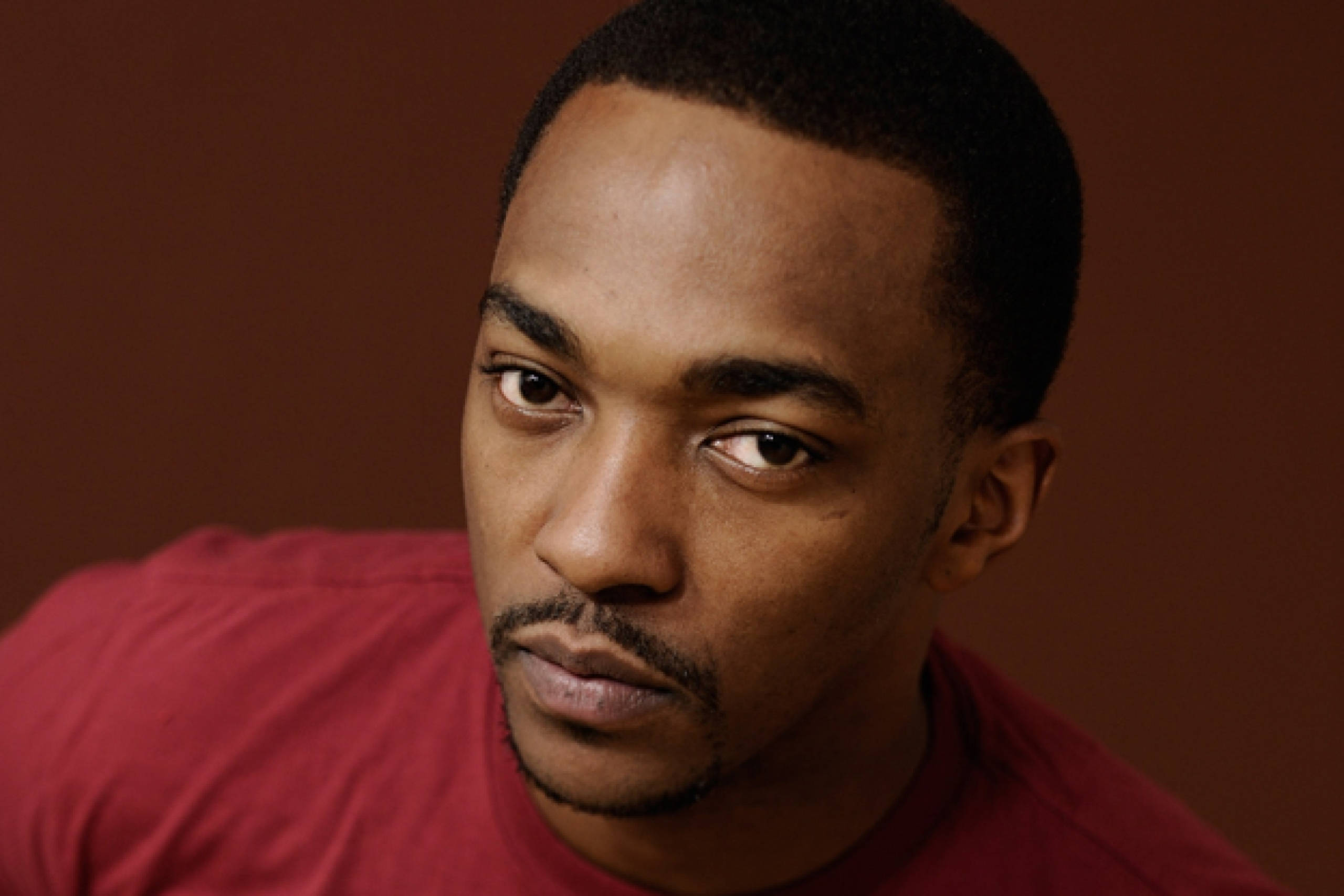 Anthony Mackie 桌布 壁紙 — 一個表情嚴肅的男人的近距離肖像，他穿著紅色襯衫，背景是溫暖的棕色。