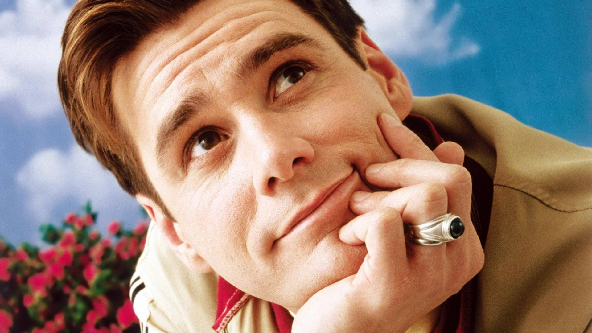 Jim Carrey 桌布 壁紙 — 一個表情深思的男人，托著下巴，背景是明亮的藍天和粉紅色的花朵。