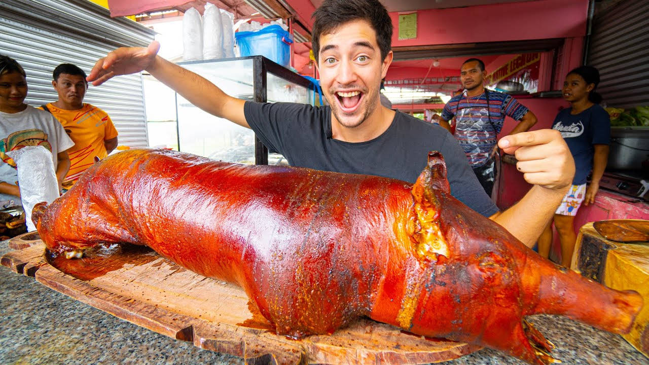 Pig 桌布 壁紙 — 一個表情興奮的男人在木製砧板旁邊自豪地比手勢，旁邊是一隻皮脆金黃的整隻烤豬。