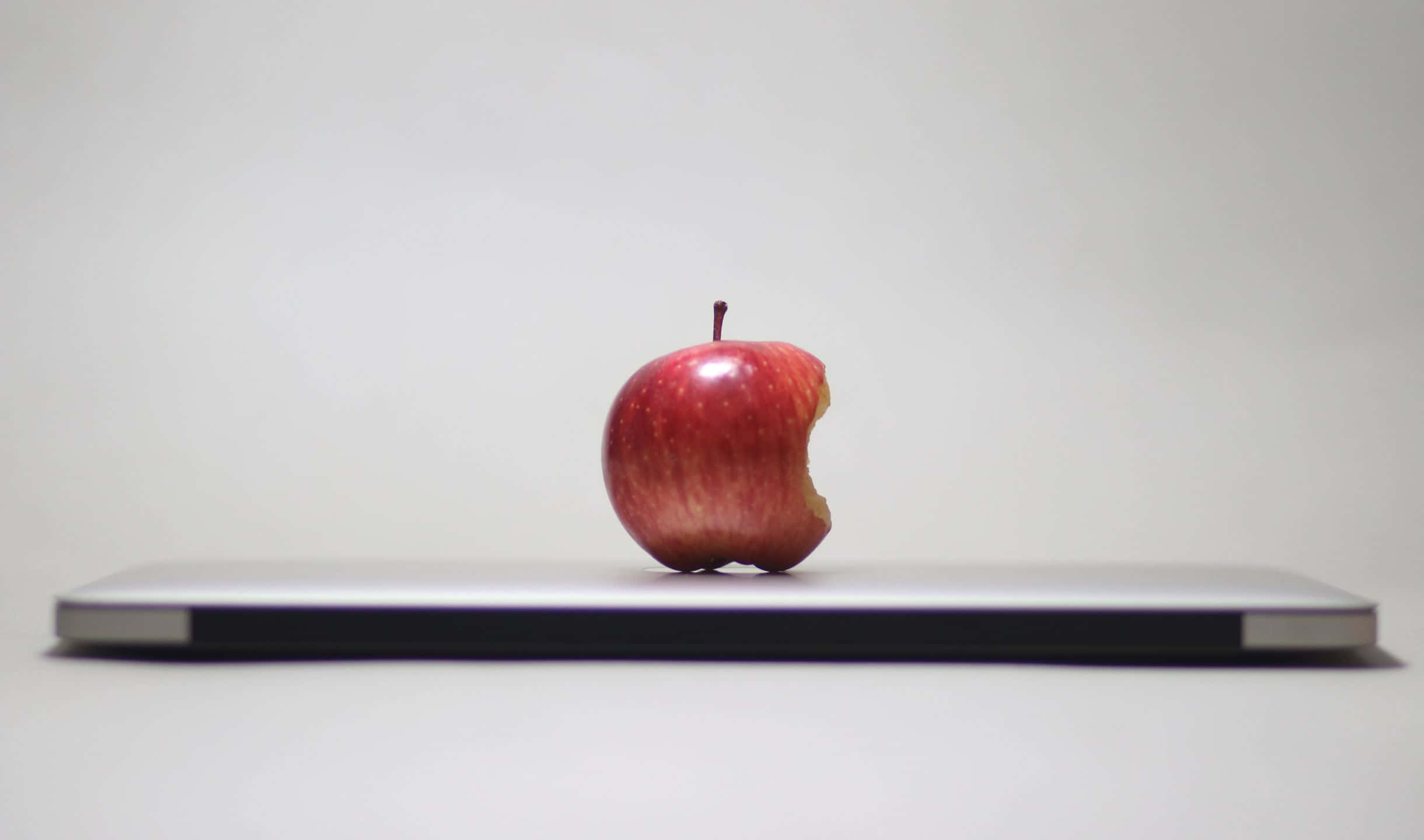 蘋果 桌布 壁紙 — 一個被咬過的紅蘋果放在關閉的銀色筆記型電腦中央，背景是純白色。