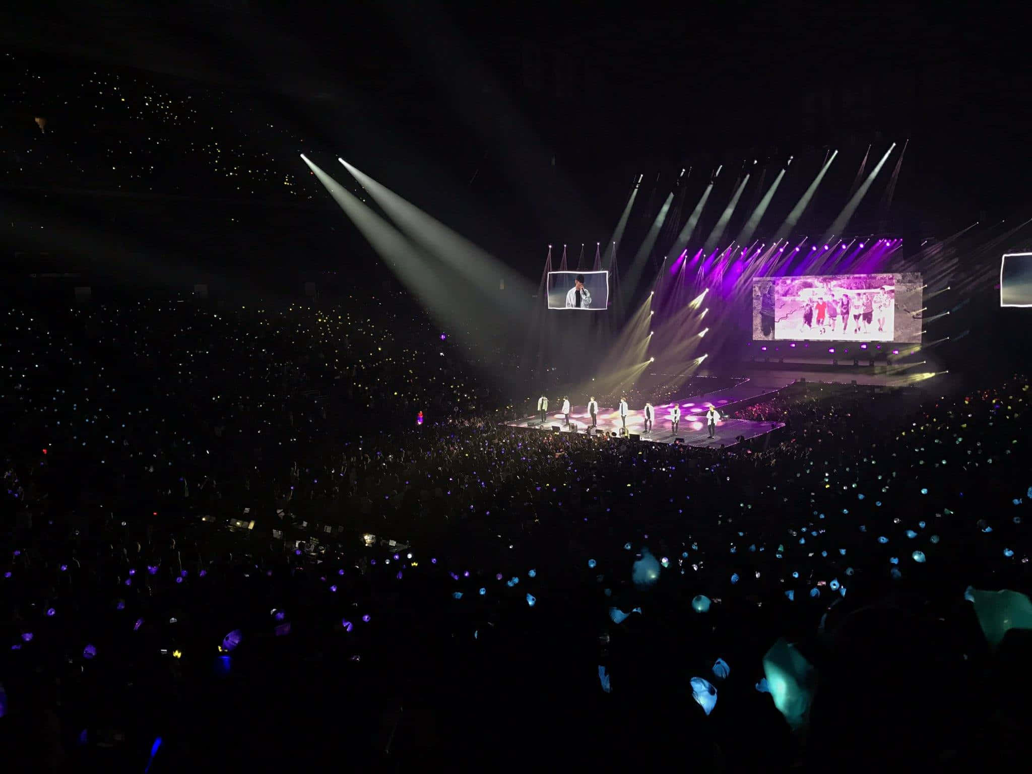 Bts Army Bomb 桌布 壁紙 — 一個被紫色和白色燈光籠罩的音樂會舞台，表演者在聚光燈下被照亮，下方是觀眾席上閃爍的藍色手機燈海。