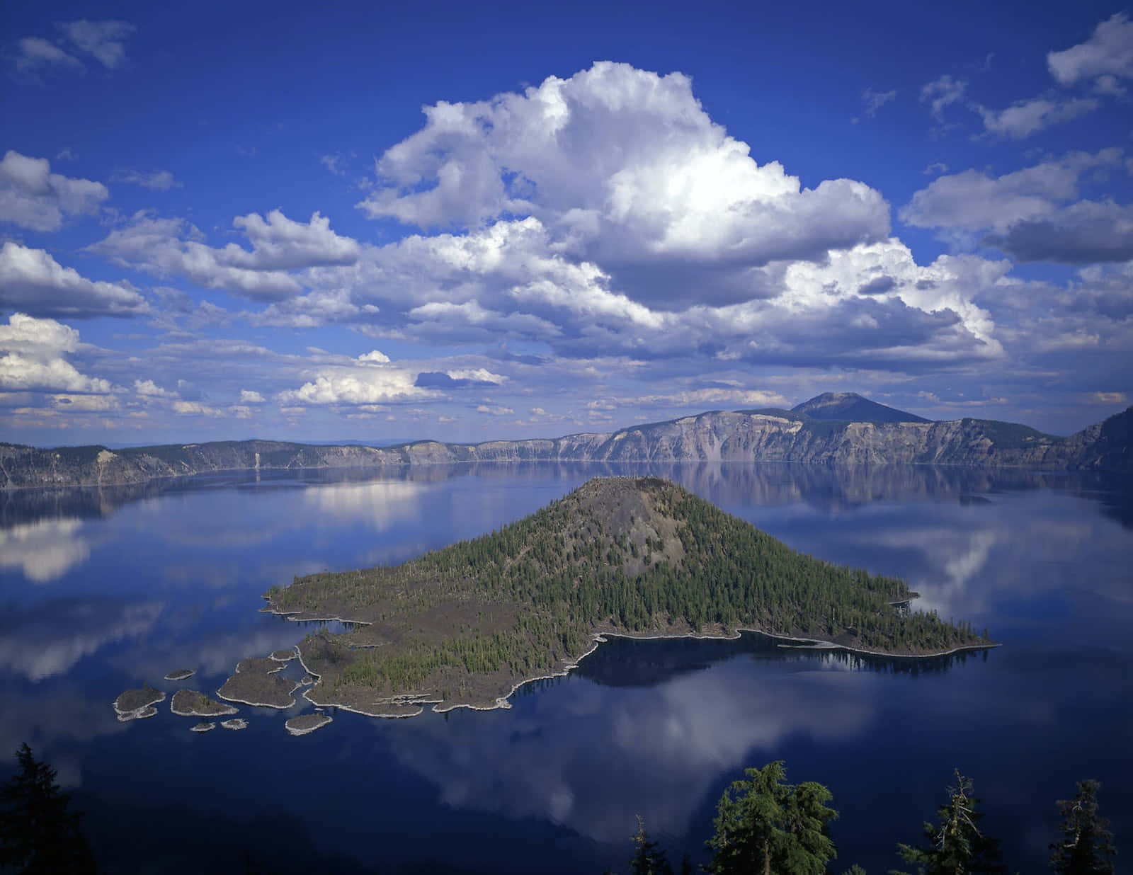 Lake 桌布 壁紙 — 一個被茂密森林覆蓋的寧靜火山島，完美地坐落於廣闊如鏡的湖泊中央，上方是明亮的藍天和蓬鬆的白雲。