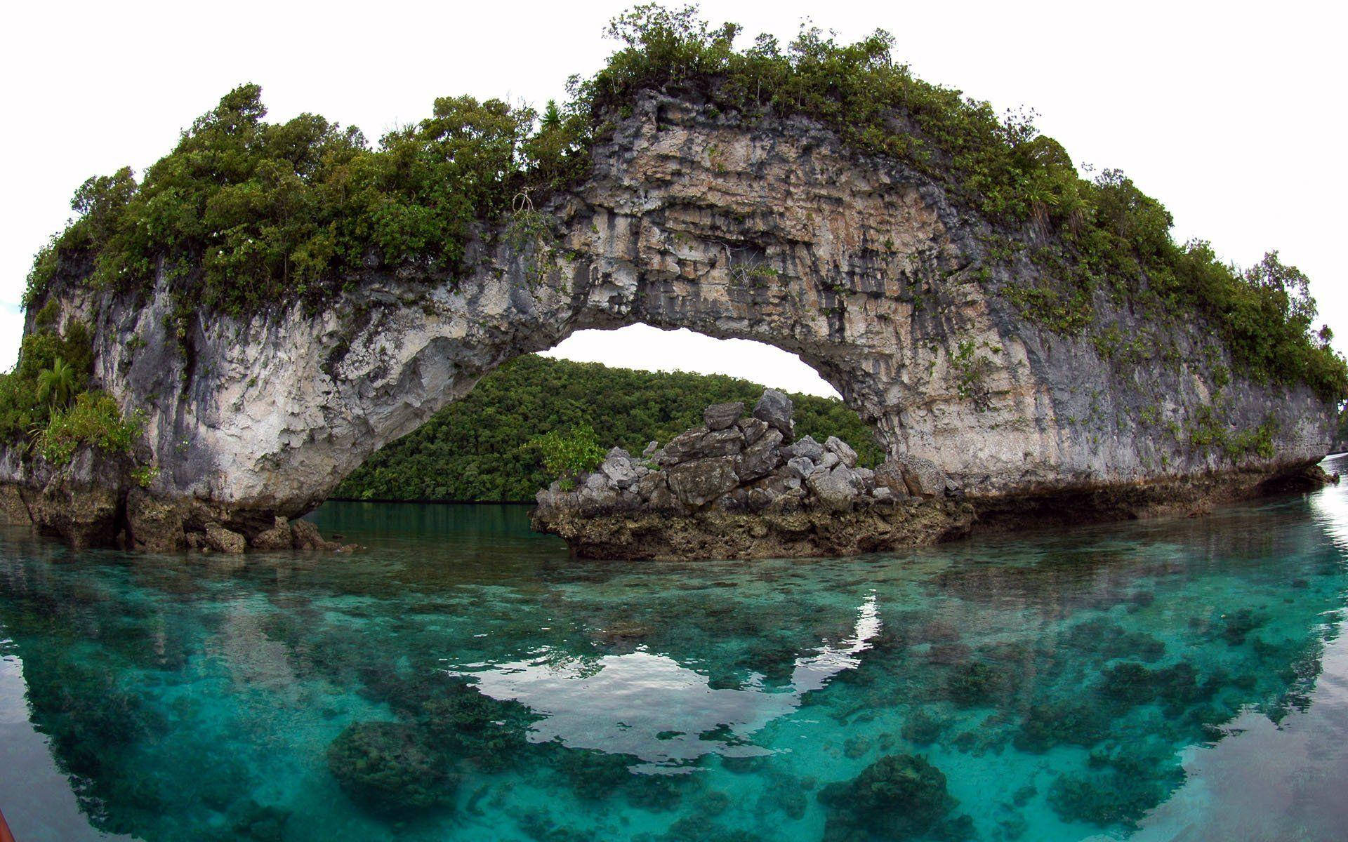 Palau 桌布 壁紙 — 一個被茂密綠植覆蓋的天然石灰岩拱門，從清澈碧綠的海水中升起，天空多雲。