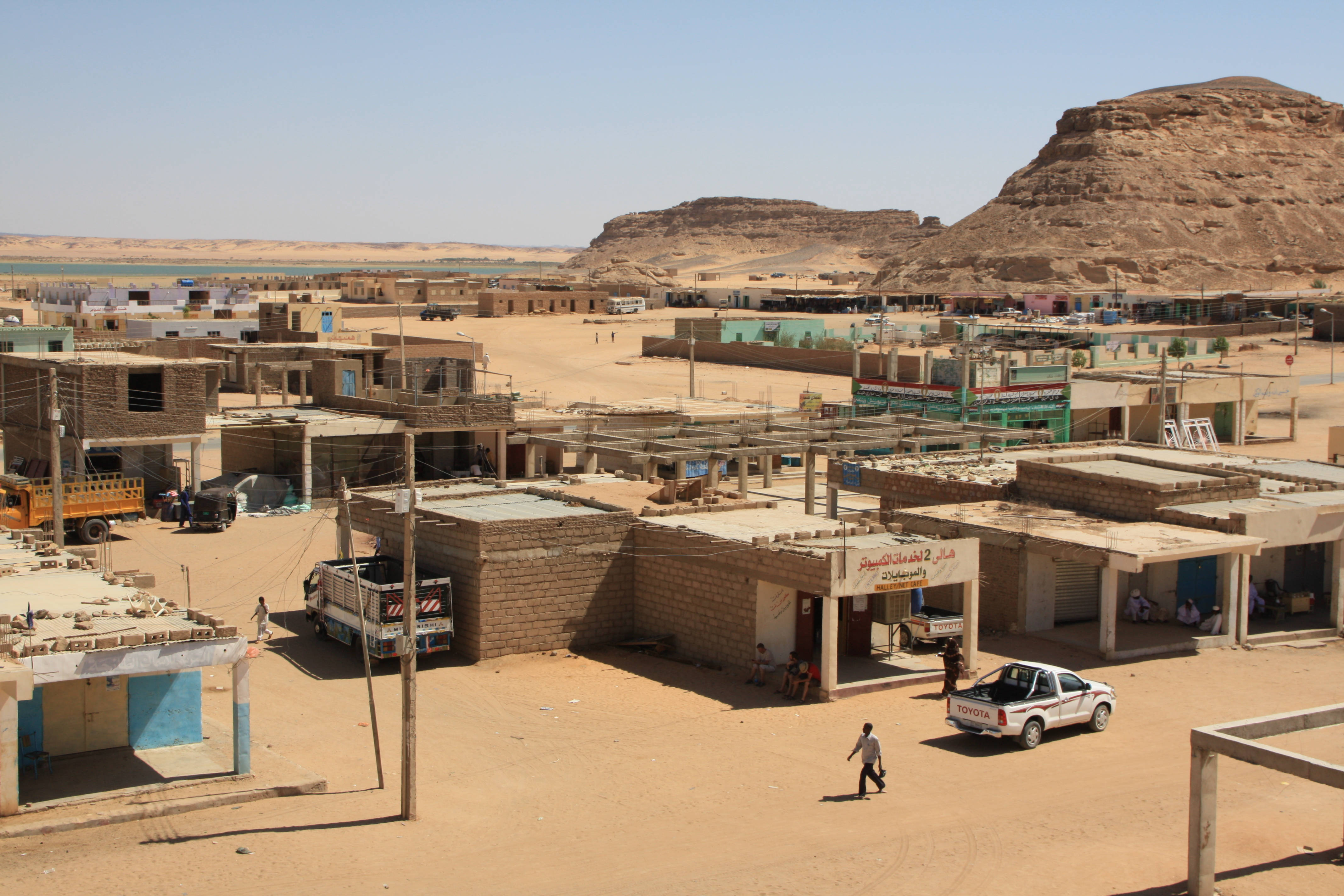 Sudan 桌布 壁紙 — 一個被陽光曬得滾燙的沙漠小鎮，有塵土飛揚的街道、低矮建築和岩石丘陵，上方是清澈的藍天。