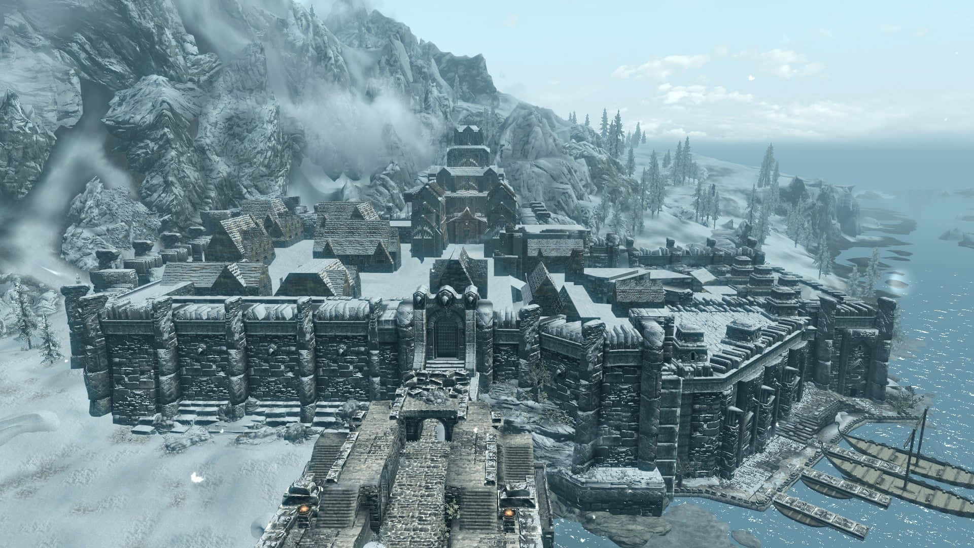 Skyrim Windhelm 桌布 壁紙 — 一個被雪覆蓋的中世紀堡壘小鎮，坐落在冰封的山脈中，旁邊是冰凍的海洋，天空蒼白。