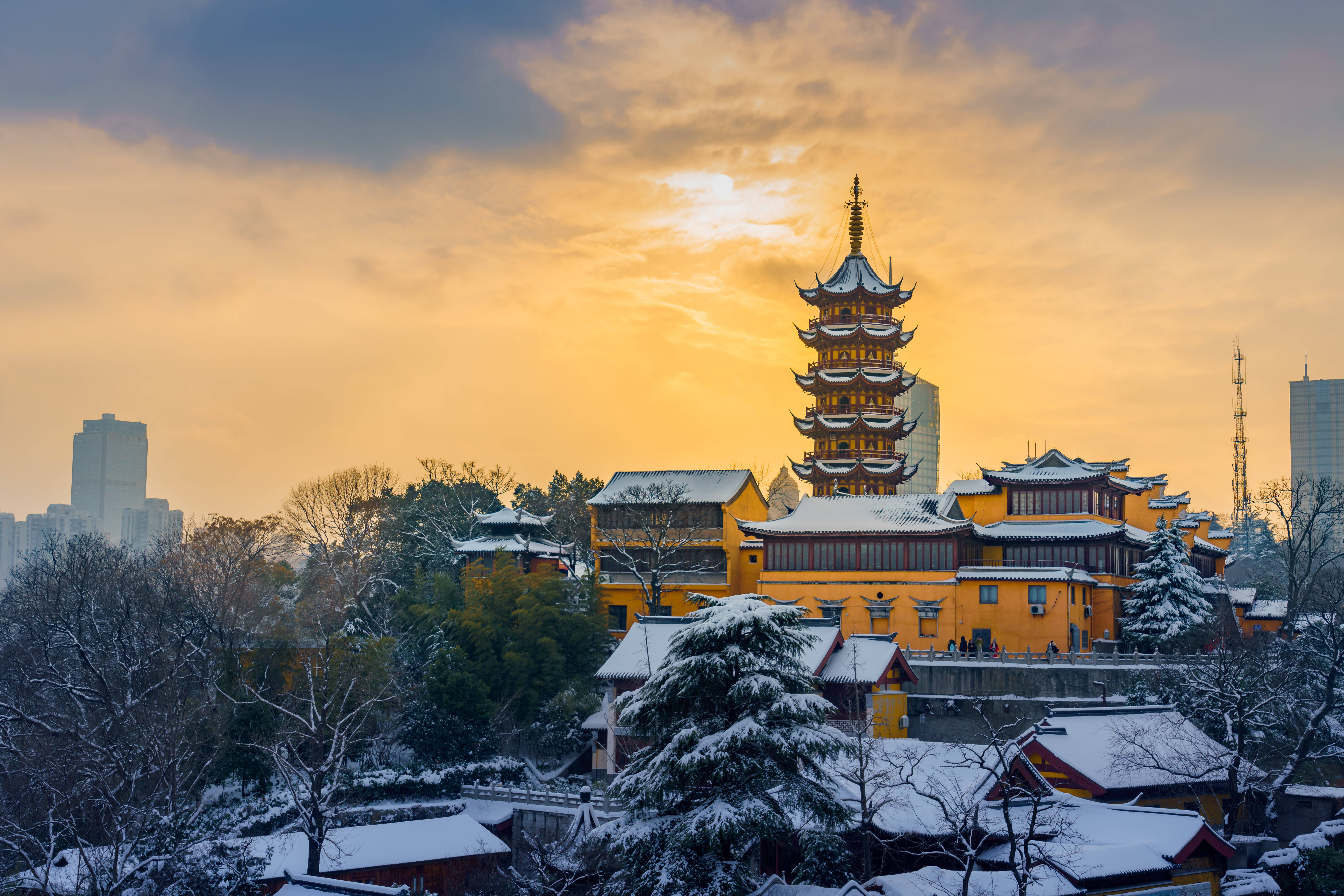 Nanjing 桌布 壁紙 — 一個被雪覆蓋的傳統寶塔和寺廟建築群，在金色夕陽天空下溫暖地發光，背景是現代摩天大樓。