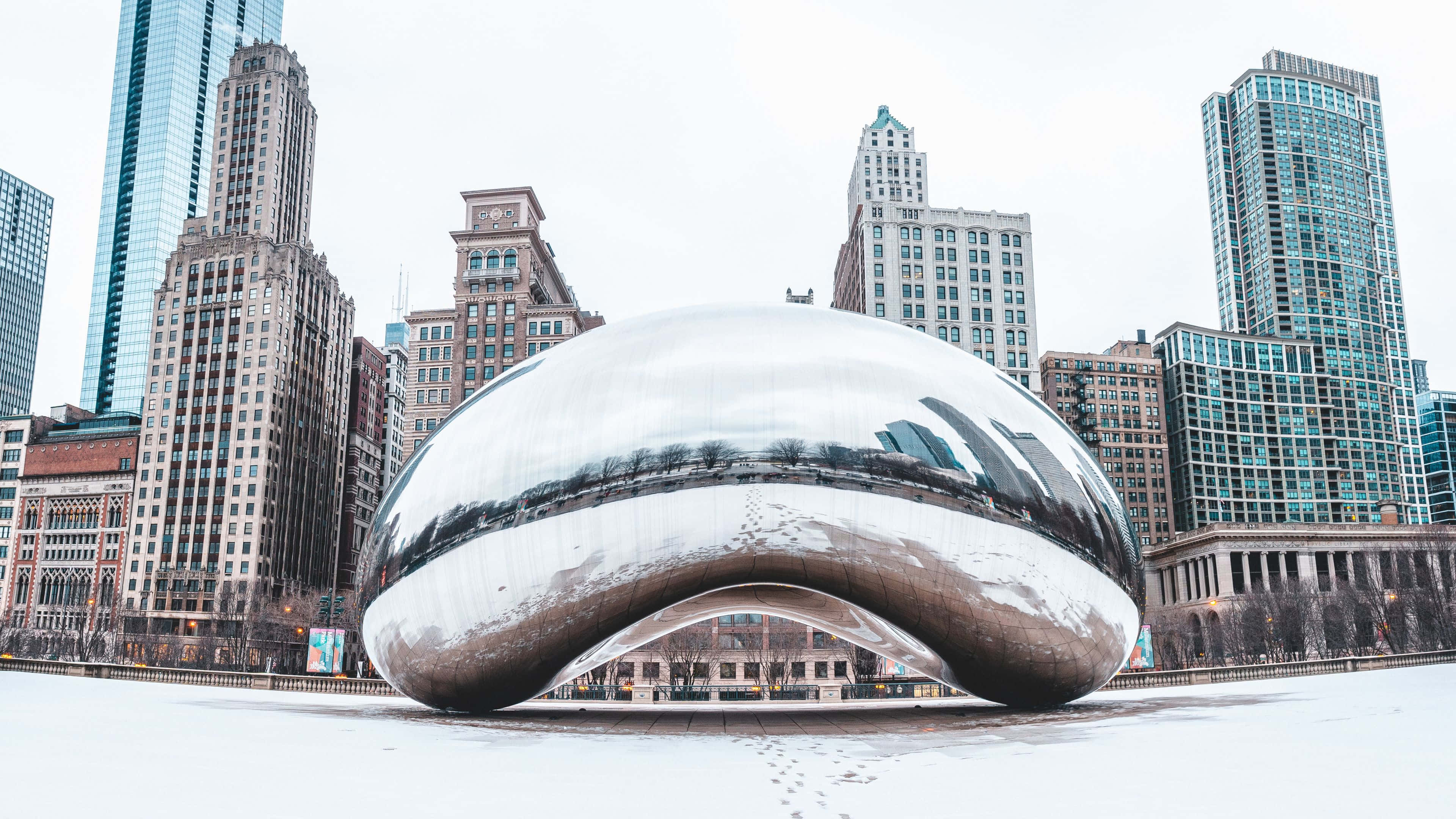 4K Ultra HD Chicago 桌布 壁紙 — 一個被雪覆蓋的反光豆形雕塑，在陰沉的冬日天空下映照著周圍的摩天大樓。