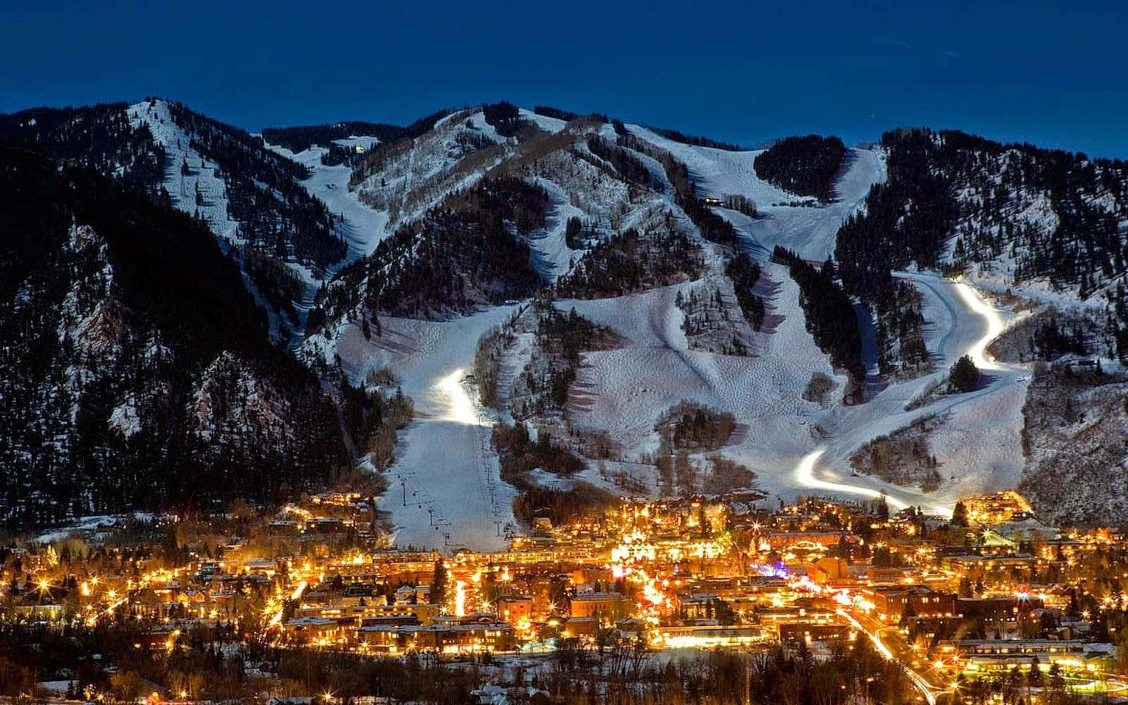 Aspen Colorado 桌布 壁紙 — 一個被雪覆蓋的山城在黃昏時分，以溫暖的燈光映照著黑暗的山峰和亮起的滑雪道。