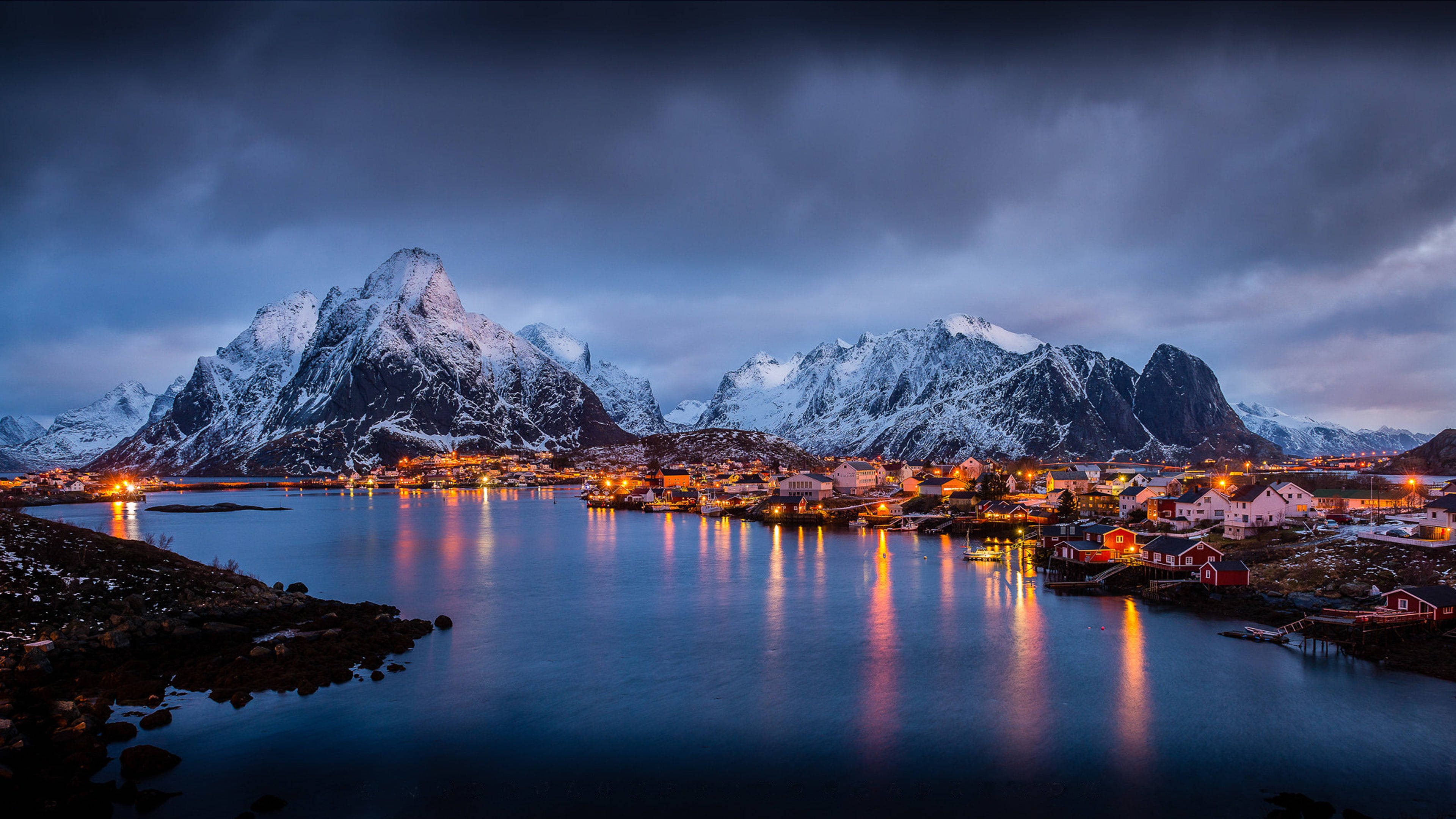 Norway 桌布 壁紙 — 一個被雪覆蓋的山村，在憂鬱的暮色天空下，溫暖的燈光映照在平靜的水面上閃爍。