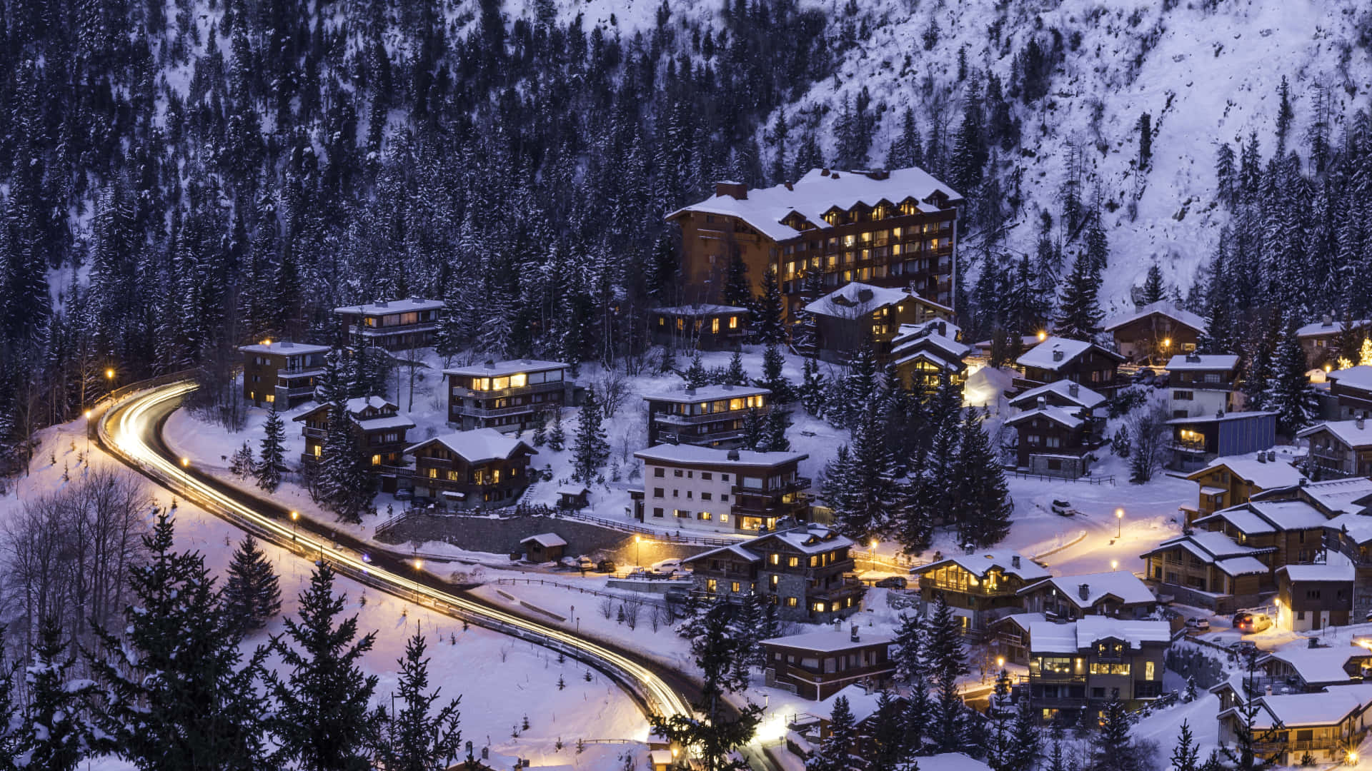 Ski Resort 桌布 壁紙 — 一個被雪覆蓋的山村，在黃昏時分，窗戶閃爍著光芒，蜿蜒的道路上佈滿了光線。
