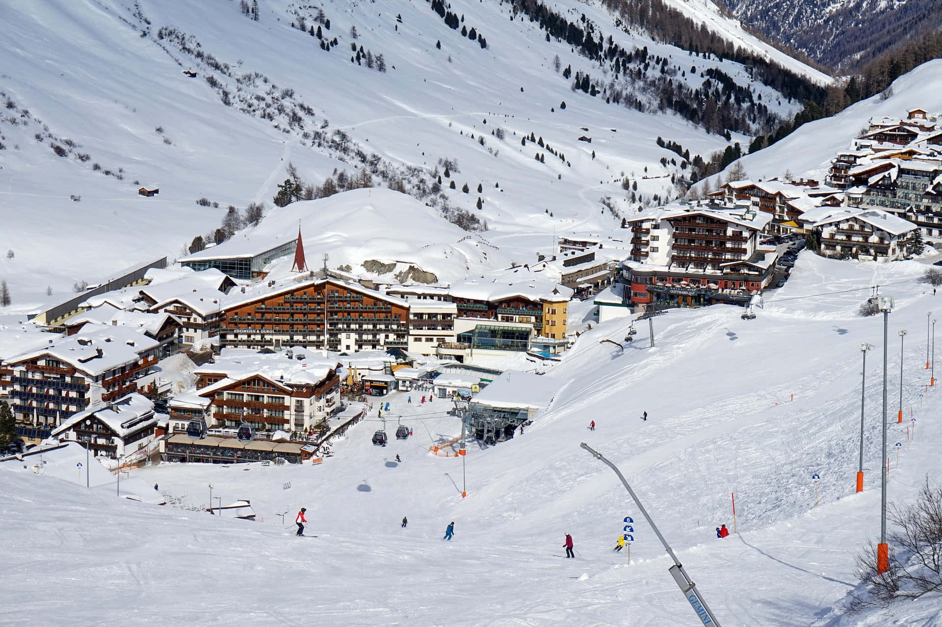 Ski Resort 桌布 壁紙 — 一個被雪覆蓋的山村，有木屋和滑雪者在明亮的冬日陽光下滑坡。