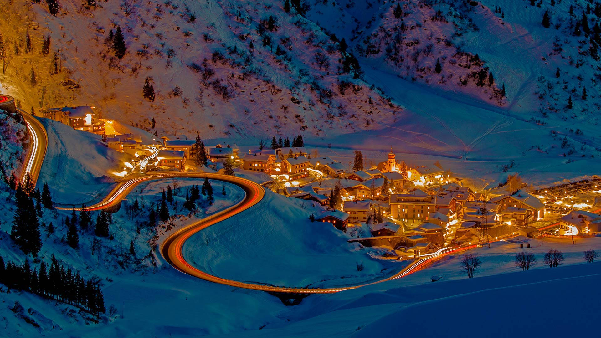 必應 桌布 壁紙 — 一個被雪覆蓋的山村在夜晚溫暖地發光，蜿蜒的道路在冰凍的景觀中留下發光的燈光軌跡。