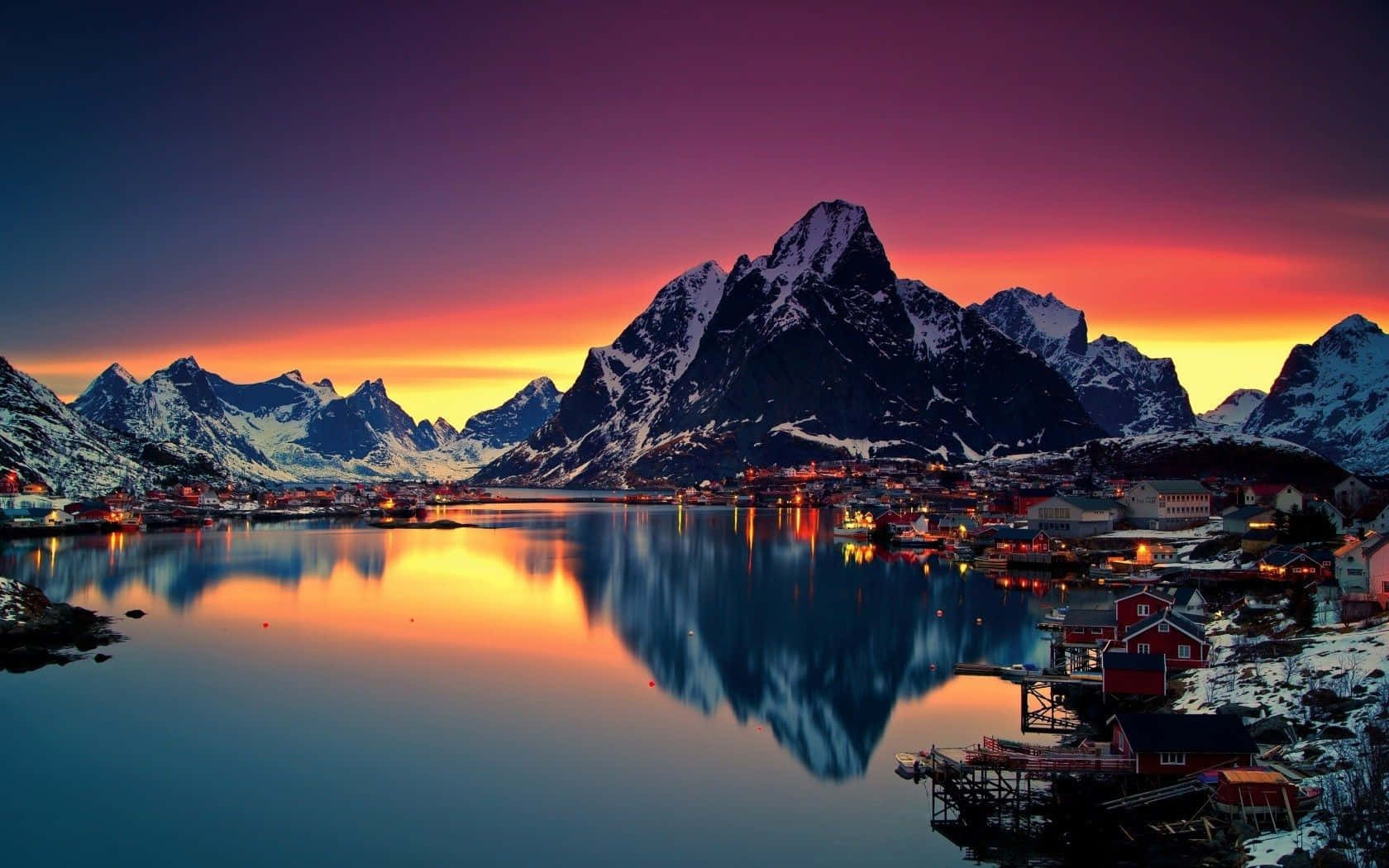 Norway 桌布 壁紙 — 一個被雪覆蓋的山村在日落時分，在平靜的水面上倒映出紫橙色天空的鮮豔色彩。
